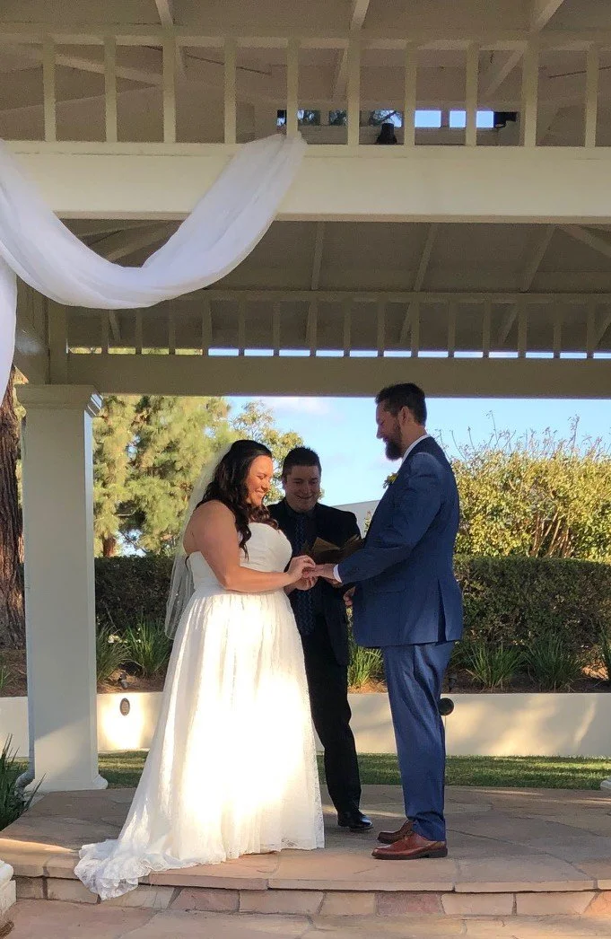A couple getting married during an outdoor ceremony, with the officiant standing between them. The bride is in a white dress, and the groom is in a blue suit. The setting appears to be a garden or park with trees and greenery in the background.