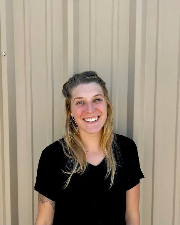A young woman with blonde hair in braids, smiling, wearing a black shirt and hoop earrings, standing in front of a beige metal wall.