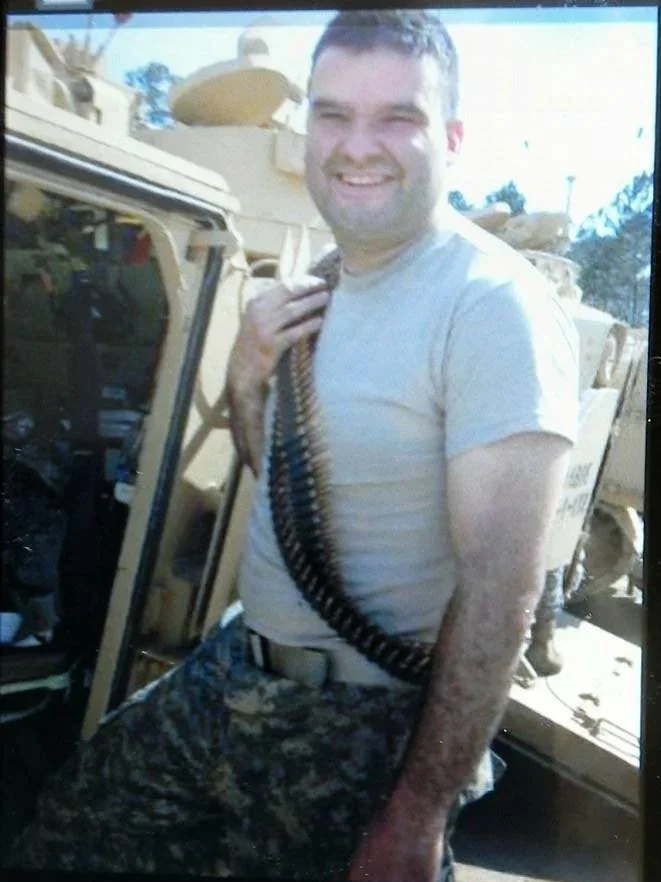 Man in military camouflage pants in front of a military vehicle, smiling, with a large ammunition belt draped over his shoulder.
