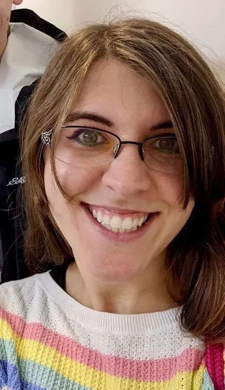 Close-up of a smiling woman with glasses and shoulder-length brown hair, wearing a colorful striped sweater.