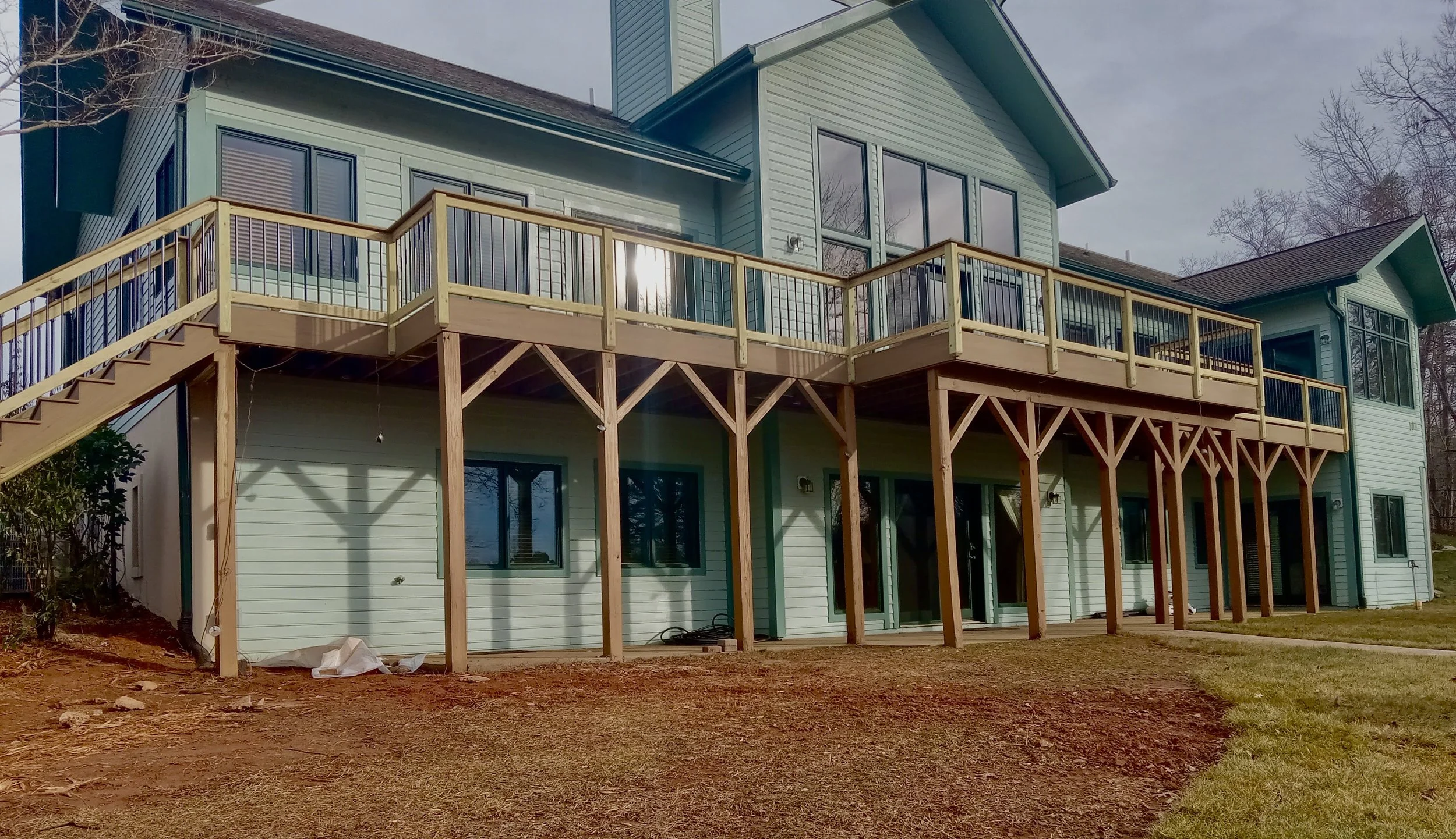 A multi-story house with a light green exterior, large windows, and a wooden deck balcony on the second floor supported by wooden beams.