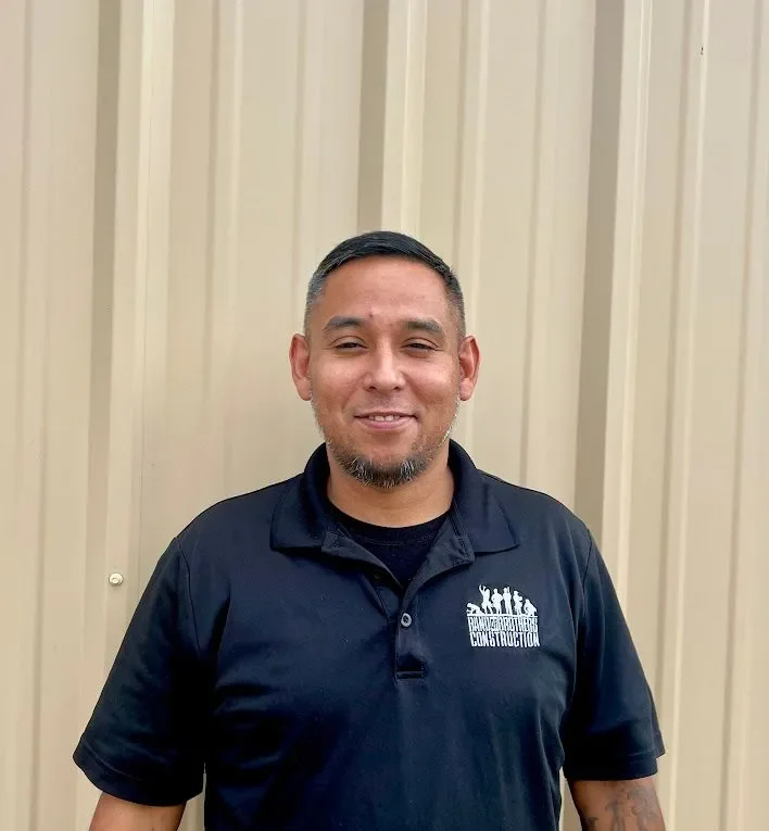 A man with short dark hair, a trimmed beard, wearing a navy blue polo shirt with a logo that reads 'The Hardworking Construction' standing in front of a beige corrugated metal wall.