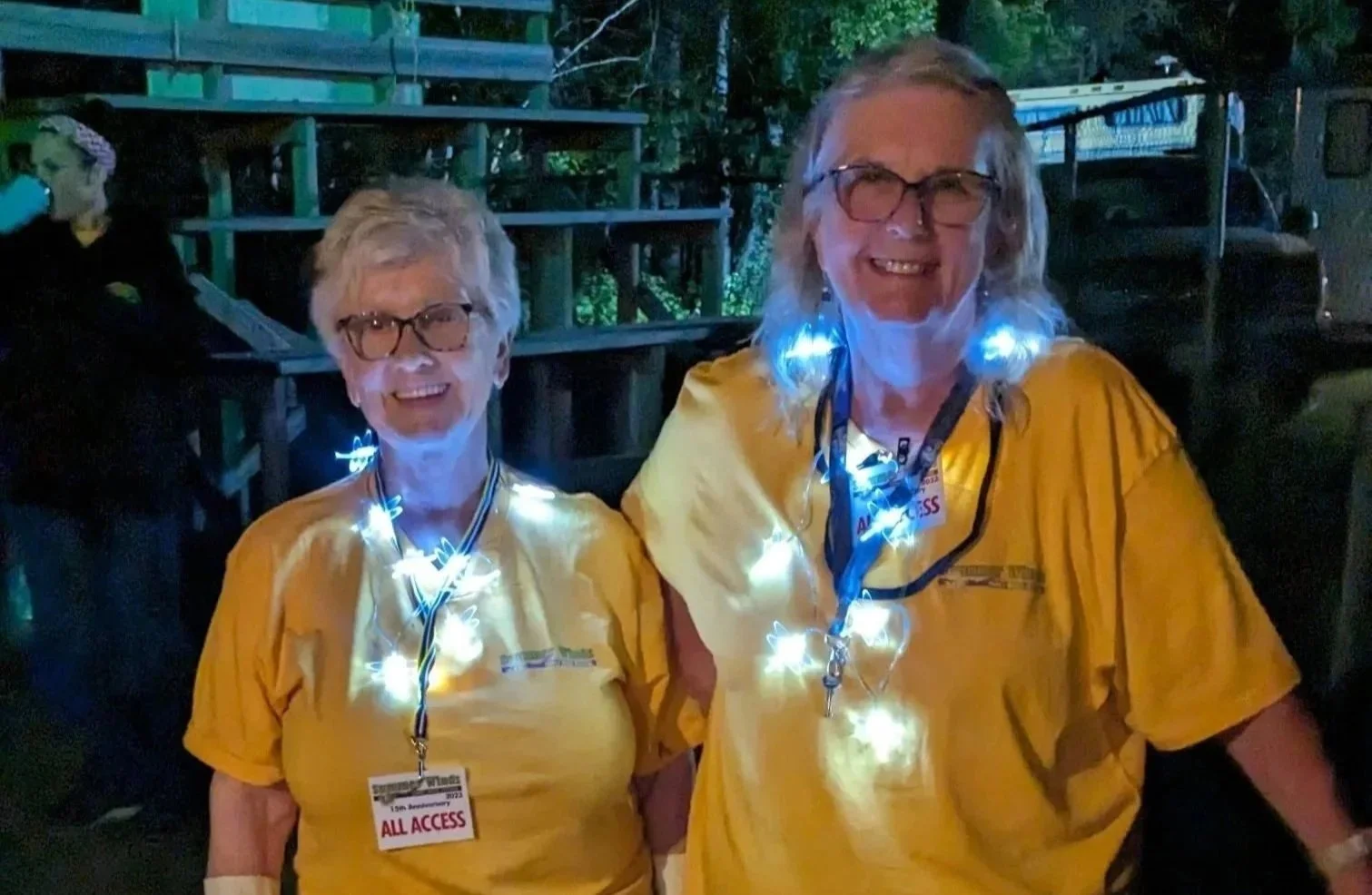 Two women smiling at night, wearing yellow shirts, glasses, and illuminated fairy lights around their necks.