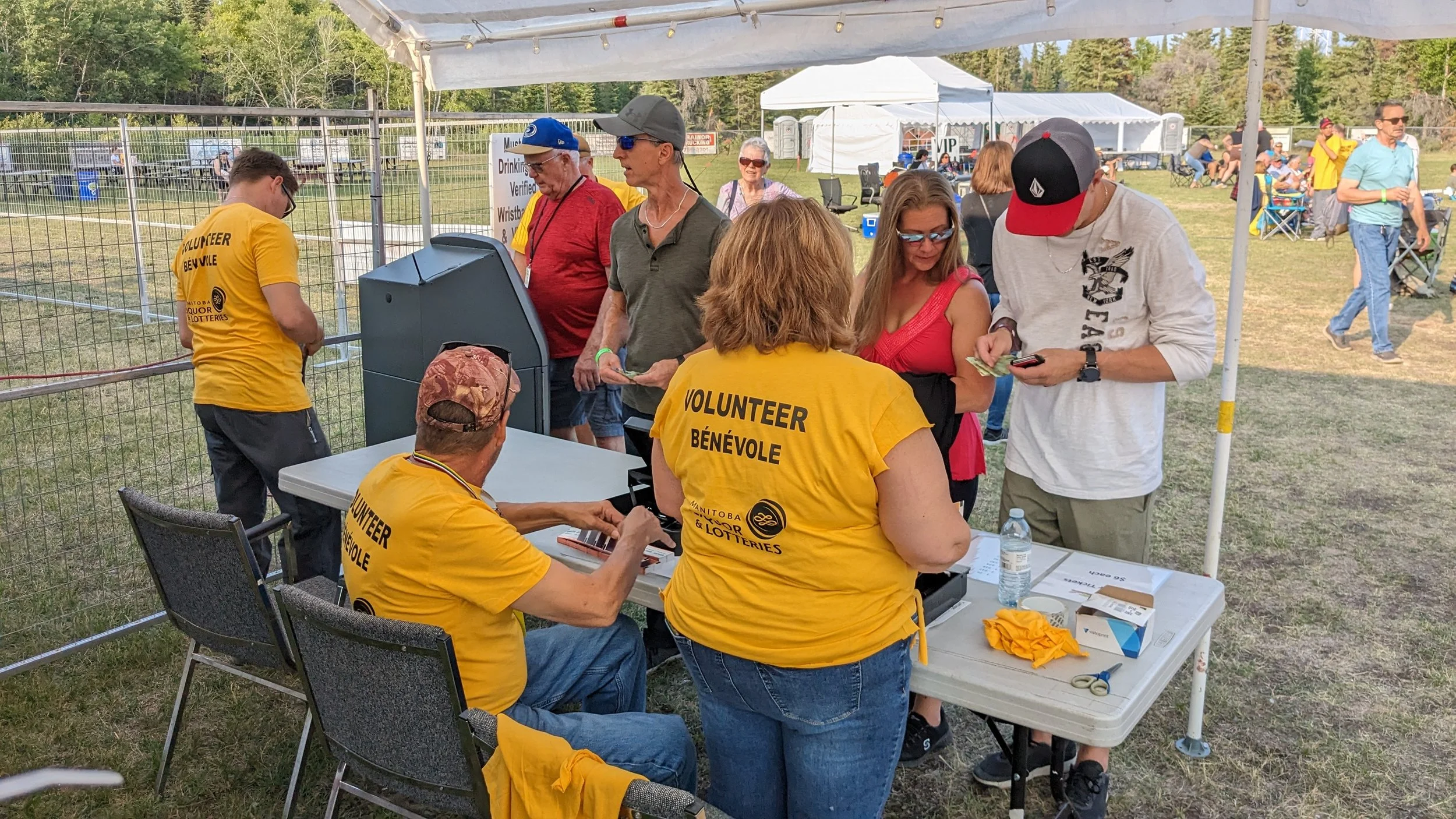 People at a registration or check-in table at an outdoor event, with some wearing yellow volunteer shirts, some on a chair and some lined up, with tents and trees in the background.