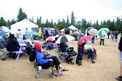 People gathered outdoors at an event with tents, umbrellas, and chairs.
