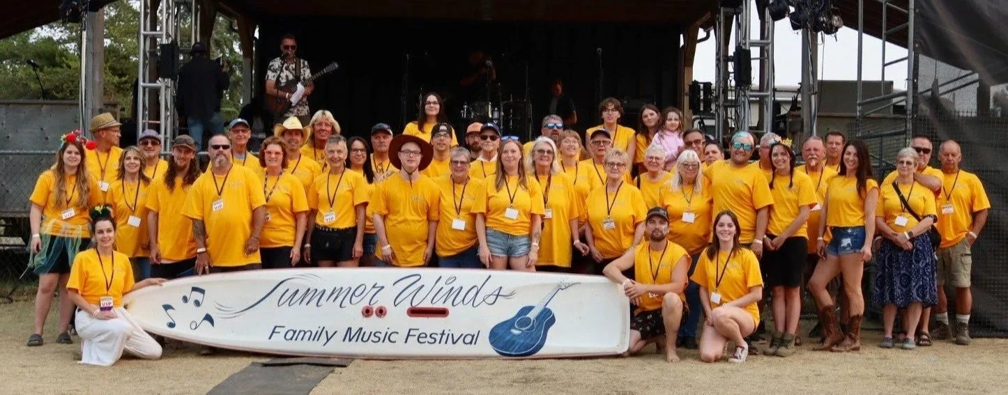 Group of people wearing yellow shirts at Summer Winds Family Music Festival, standing on stage with a banner and a live band performing in the background.