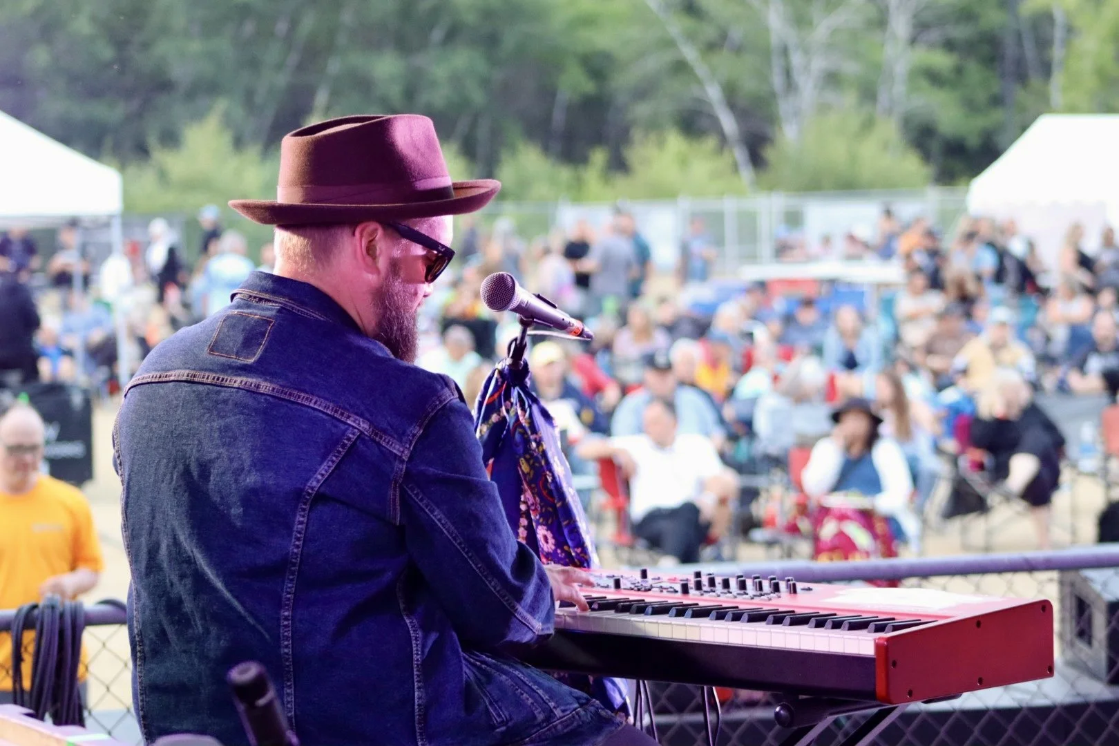 A man with a beard, wearing sunglasses, a fedora, and a denim jacket playing a red keyboard on stage during an outdoor event, with a crowd of people sitting and standing in the background.