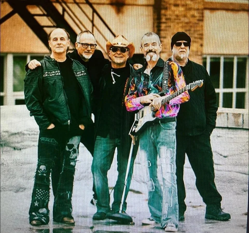 A group of five men standing together outdoors, one holding an electric guitar, with a brick building and windows in the background.
