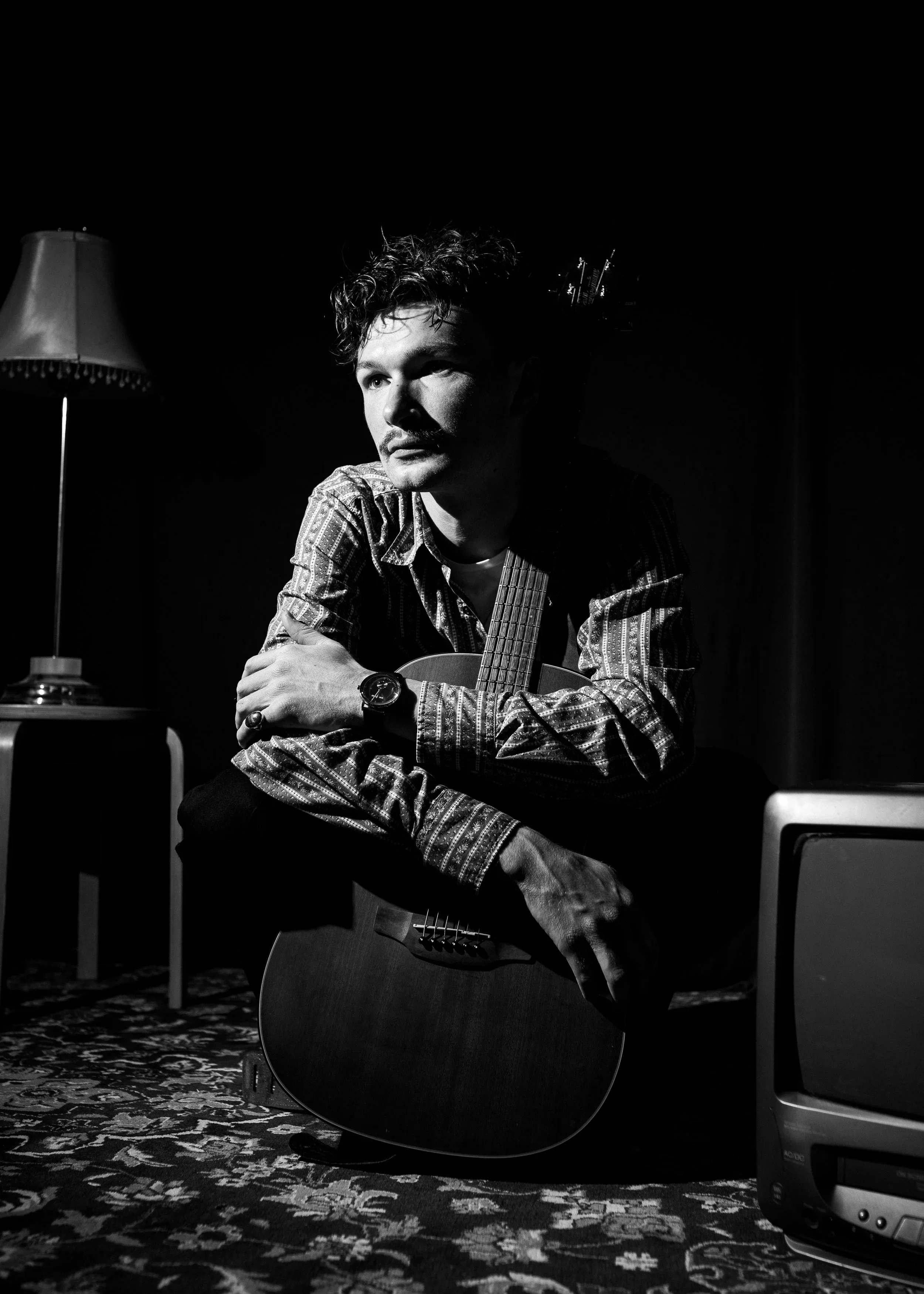 Black and white photo of a man with curly hair sitting in a room, holding a guitar. There is a lamp on a side table on the left and a vintage-style rug on the floor. The room is dimly lit.