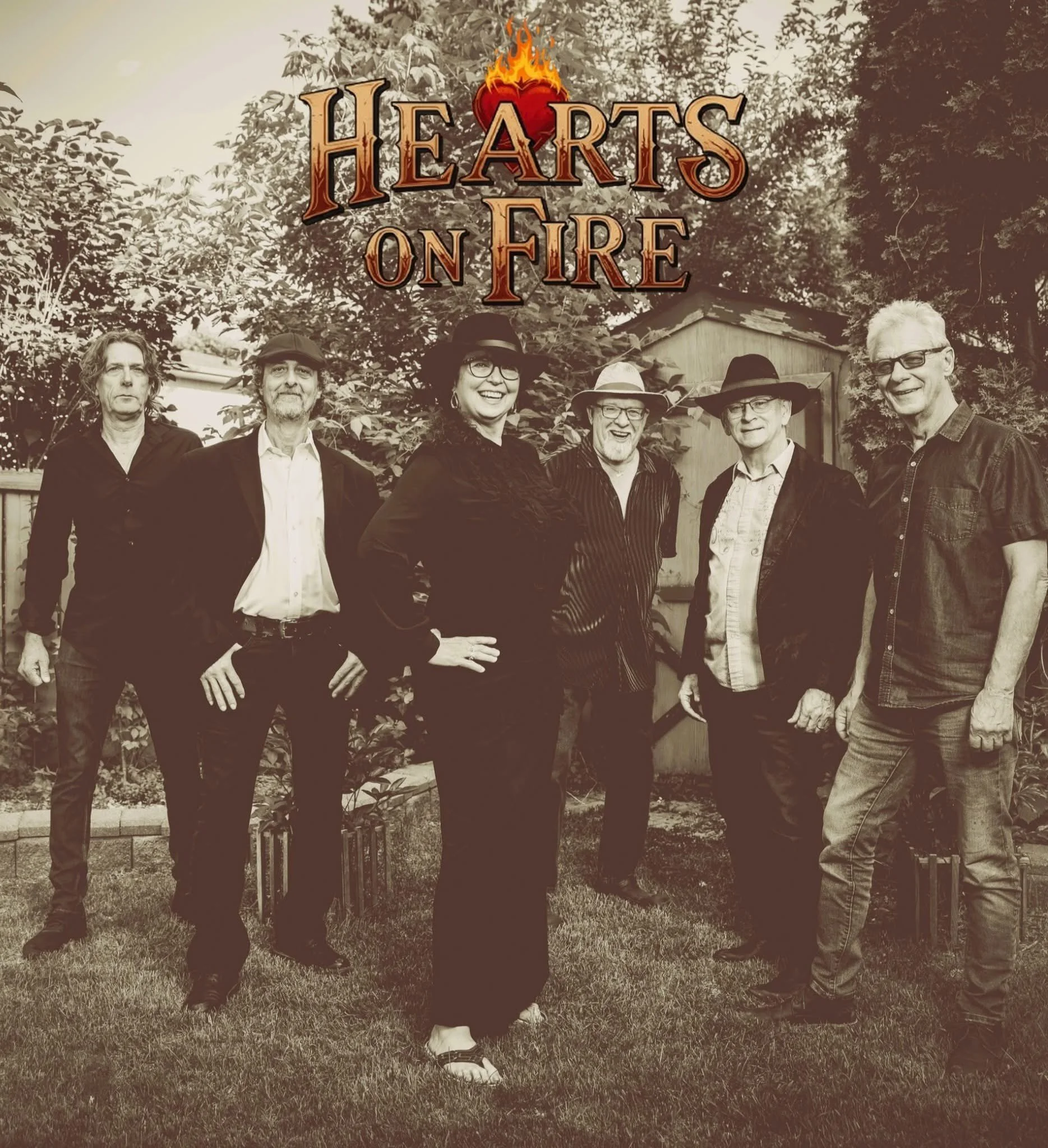 A group of six adults standing outdoors on grass with trees and a small shed in the background. The image is in black and white with a sepia tone. Overlaid is the text 'Hearts on Fire' with a flaming heart icon above it.