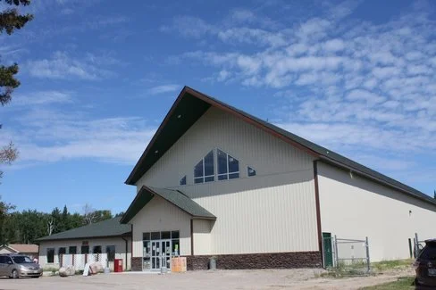 A large modern building with a sloped roof and triangular windows, surrounded by parked cars and a chain-link fence under a partly cloudy sky.