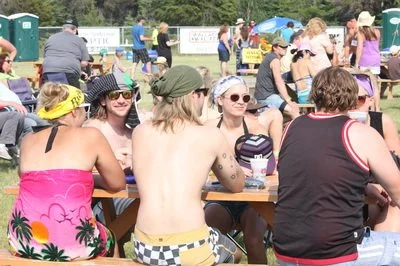 Group of people sitting at a picnic table outdoors at a festival or event, with others standing and walking in the background.