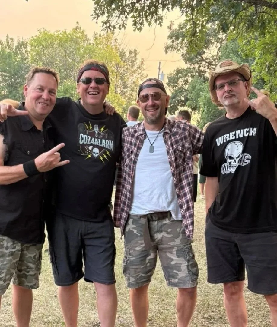 Four men posing outdoors in casual clothing, some wearing sunglasses and military-style camouflage shorts, with trees and a partly cloudy sky in the background.
