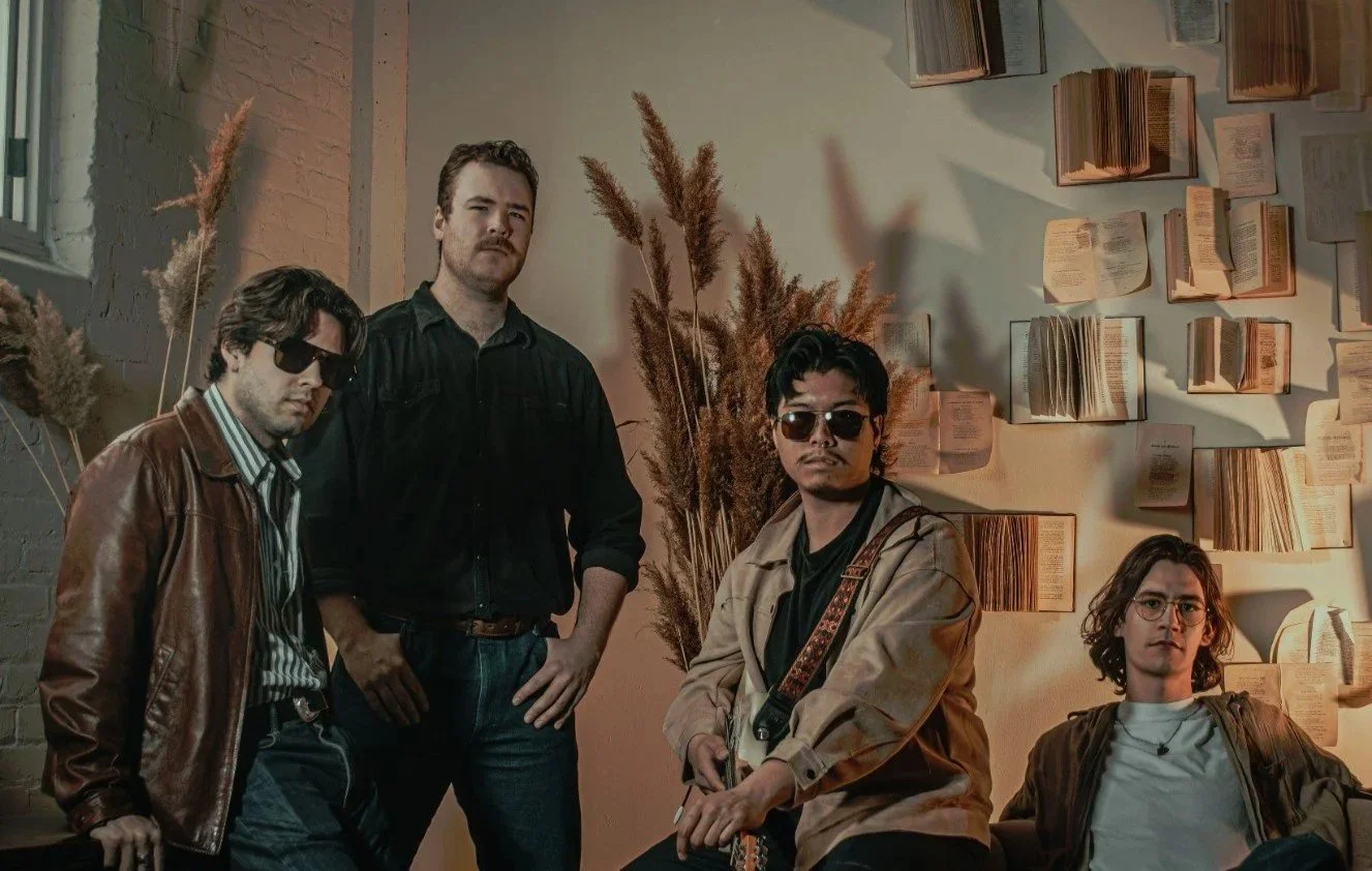 Five men posing indoors with books on the wall and plants in the background. The men wear casual clothing, and some wear sunglasses.