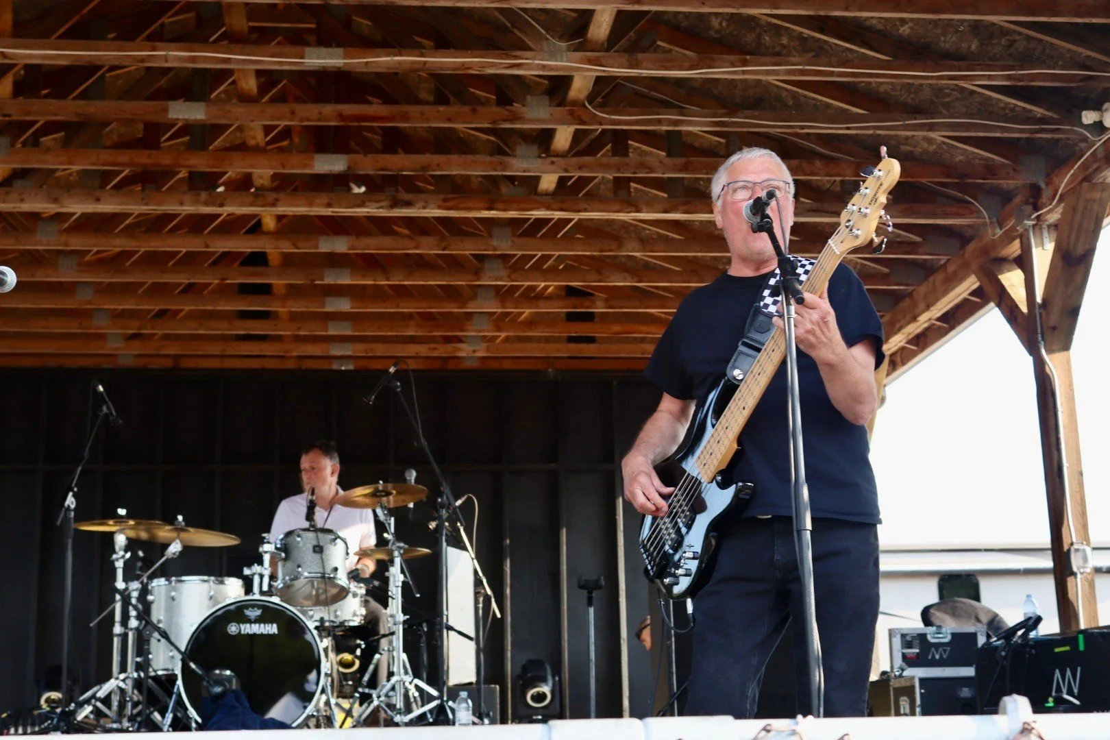 An older man with gray hair and glasses playing a bass guitar and singing into a microphone on stage, with a drummer playing a Yamaha drum set in the background, under a wooden roof structure.
