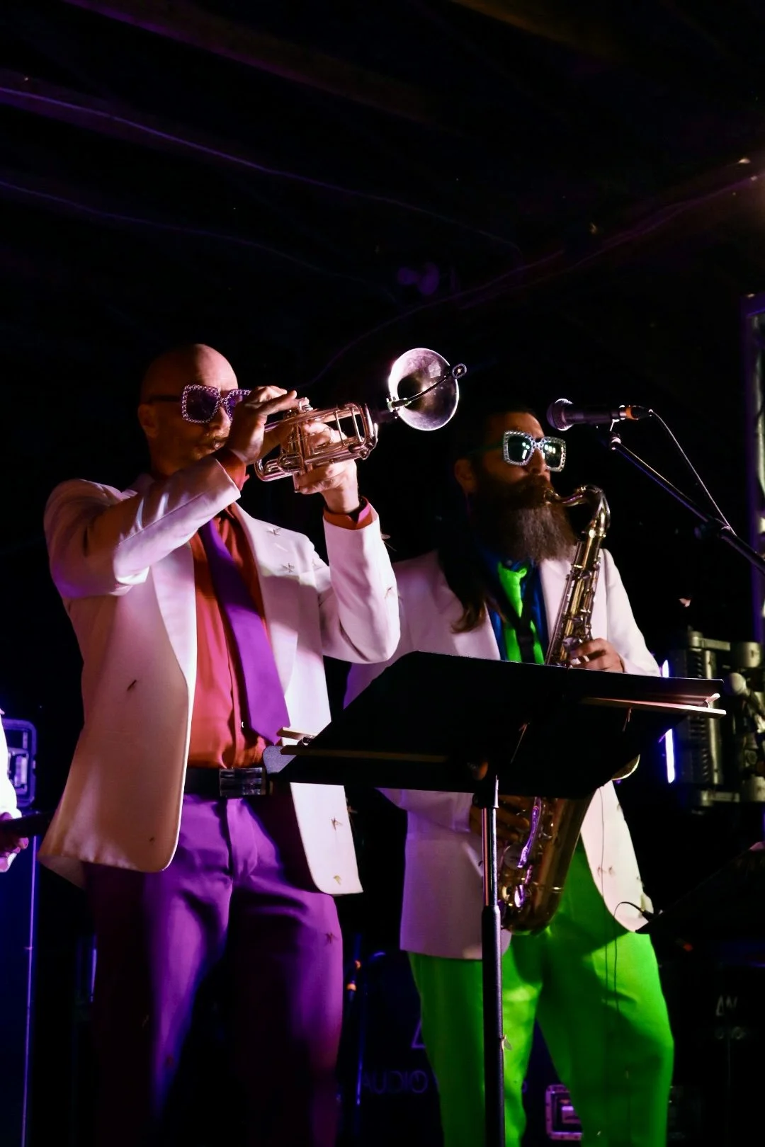 Two musicians performing on stage, one playing a trumpet and the other a saxophone, both wearing white suits and oversized sunglasses with rhinestones, illuminated by colorful stage lighting.