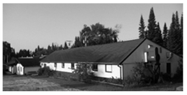 Black and white photo of a long, single-story building with a roof, trees, and a clear sky in the background.