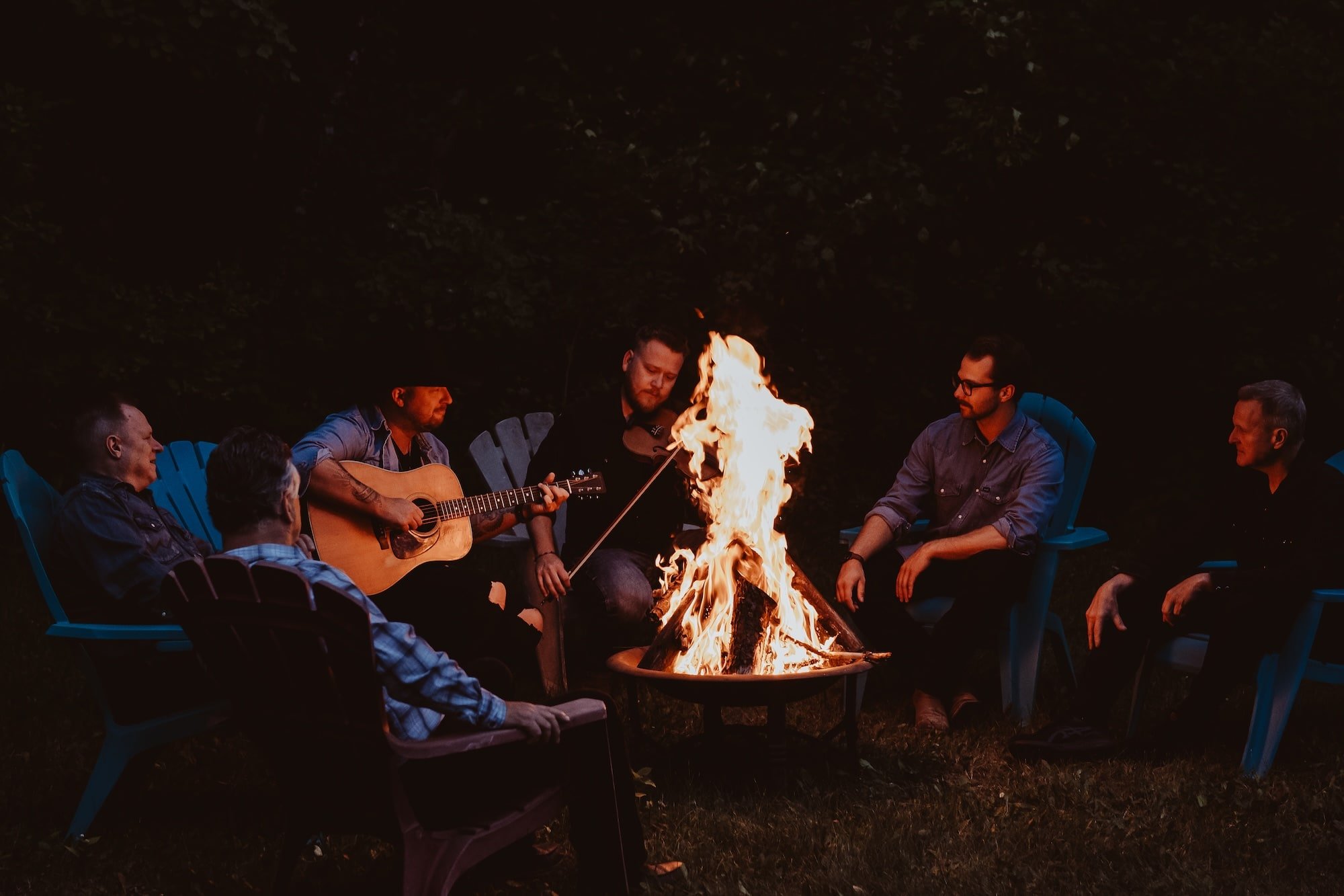 Group of people sitting in chairs around a campfire at night, with a man playing guitar and another person holding a lit match or torch near the fire.