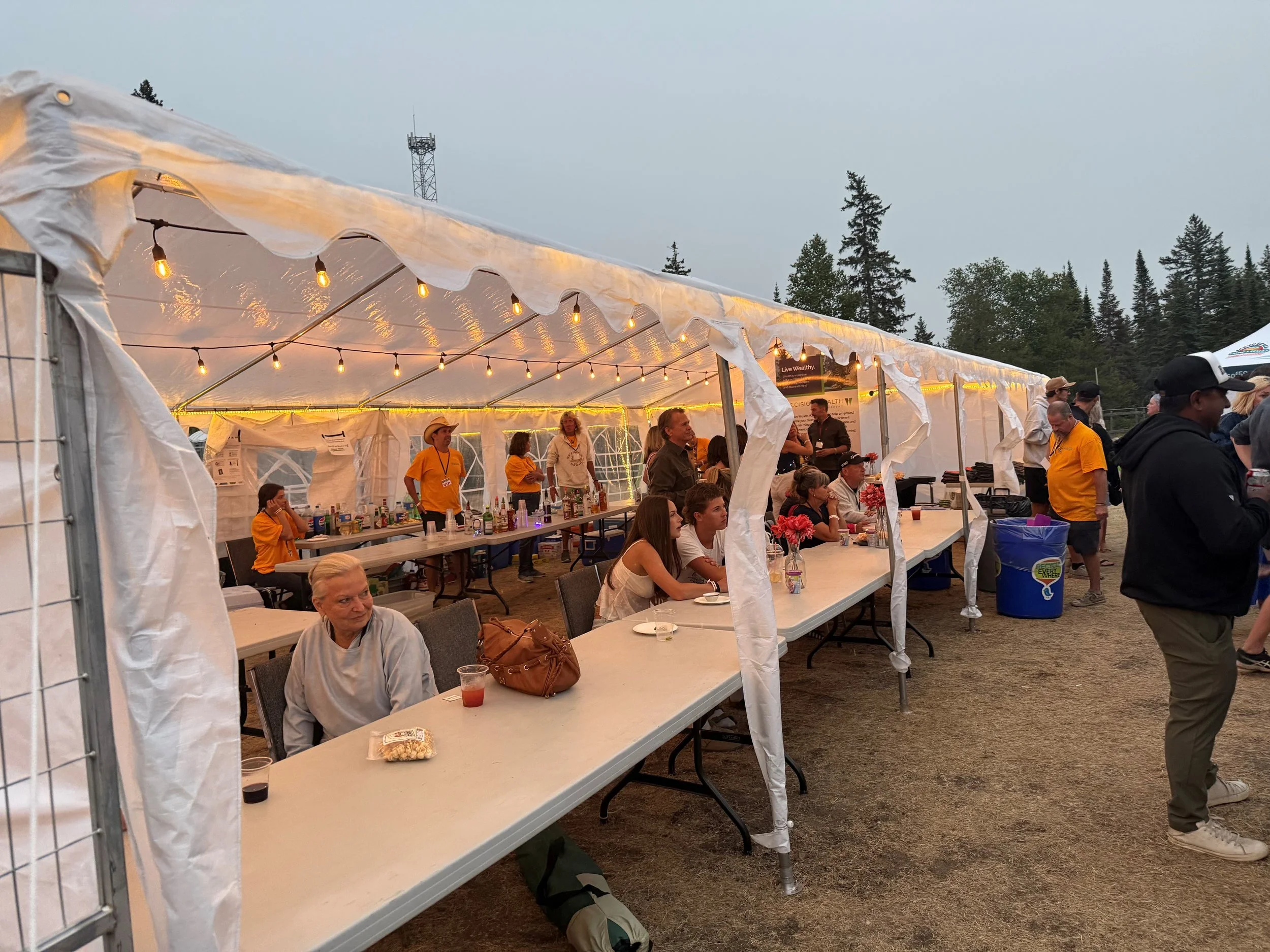 People gathered under a white event tent decorated with string lights, seated at tables with drinks and food, in an outdoor setting with trees and a cloudy sky in the background.