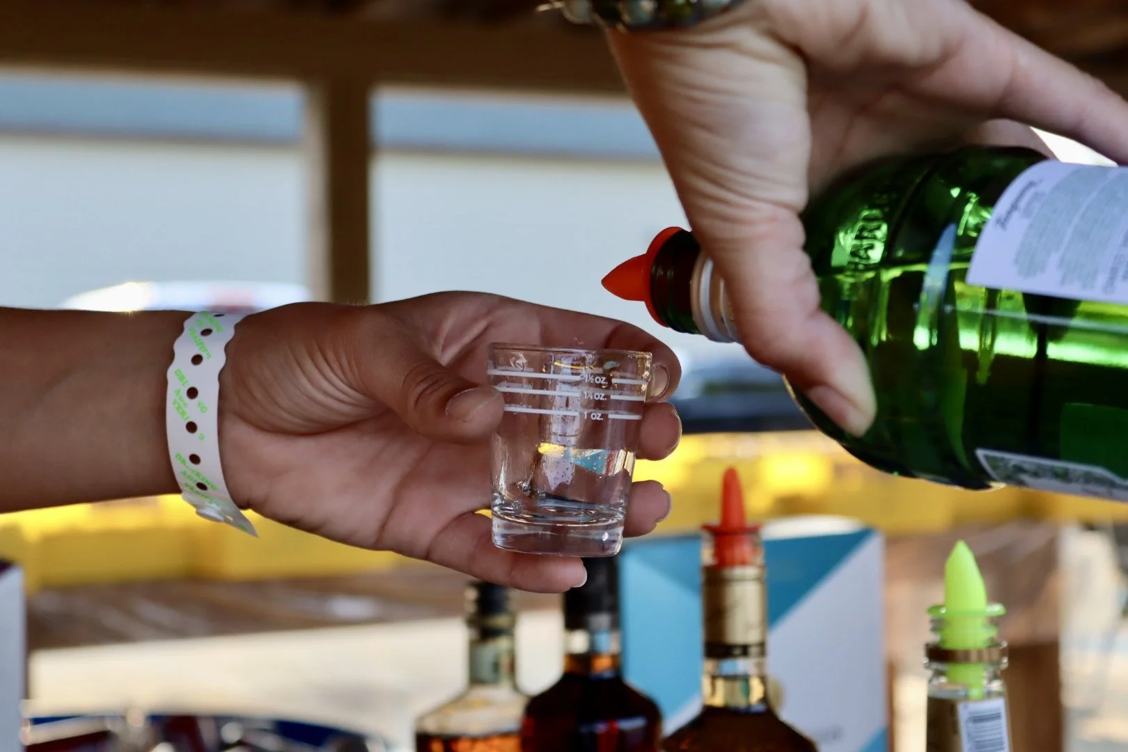 Person pouring clear liquid from a green bottle into a small glass measuring cup held by another person, with syringes and bottles in the background.