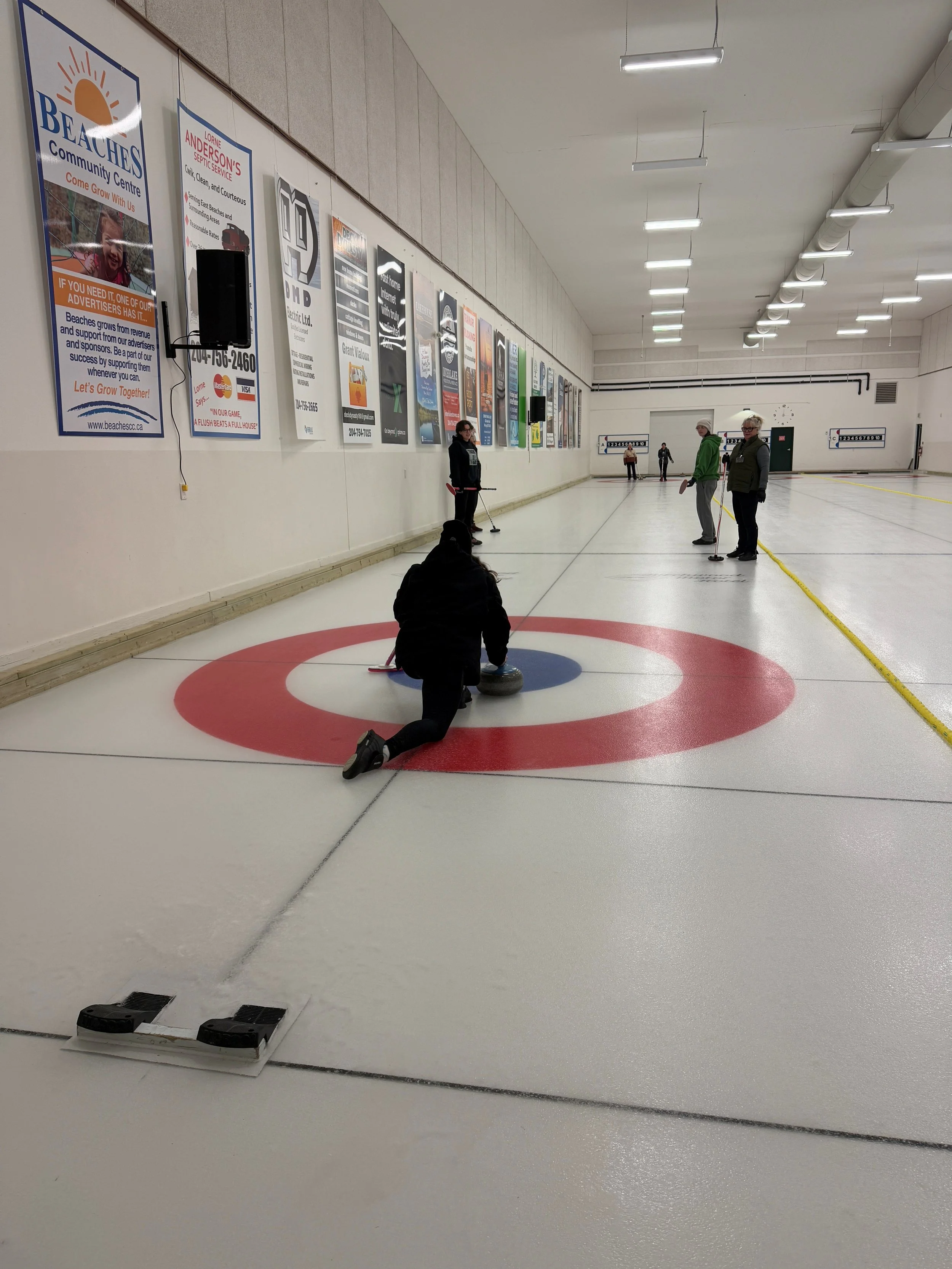 An indoor curling rink with several people preparing to play. One person is kneeling on the ice, holding a curling stone, while others stand nearby holding brooms. The rink features curling targets on the ice and posters on the wall.