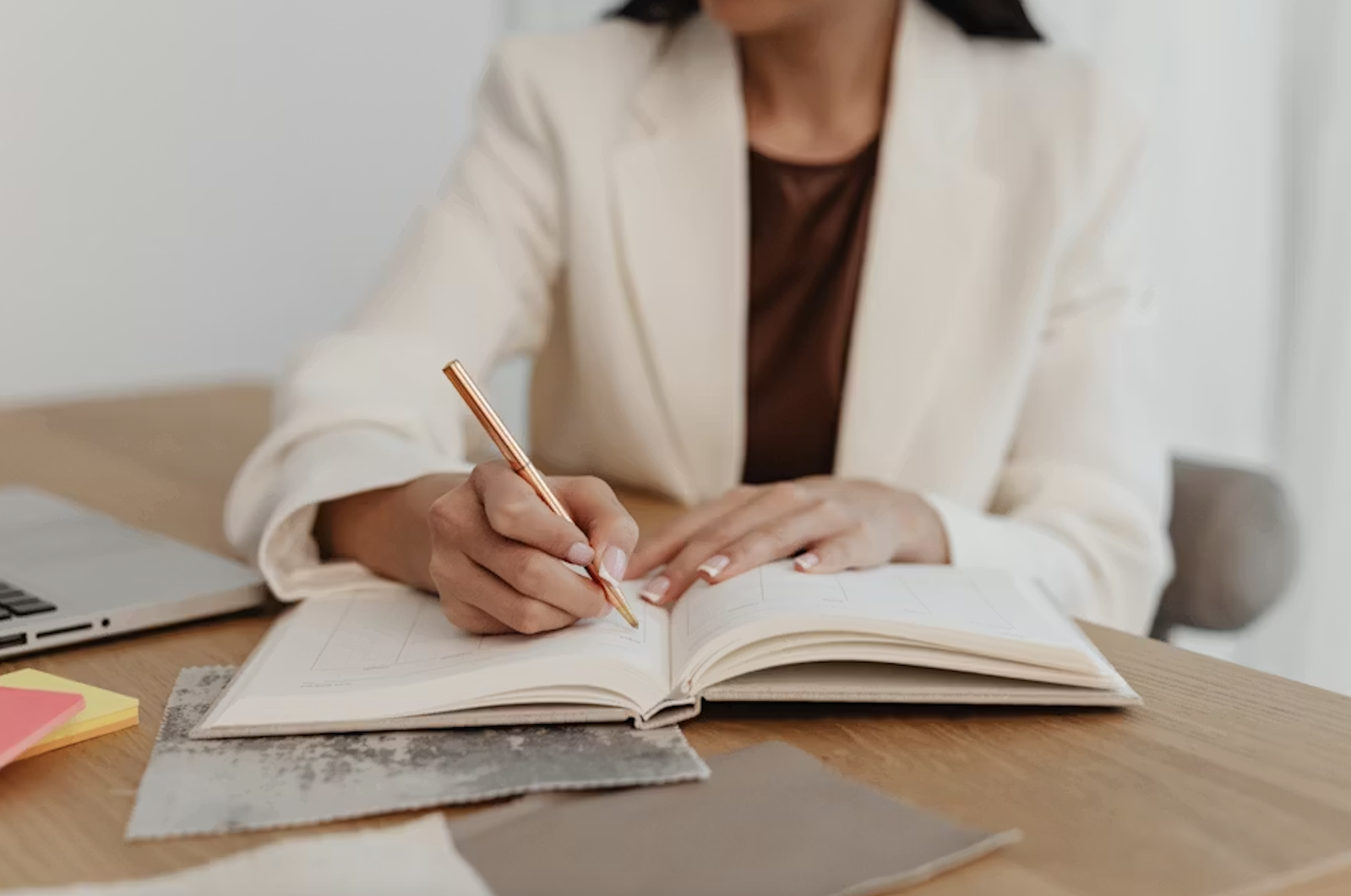 A woman in a beige blazer and dark top is writing in an open planner at a wooden desk, with sticky notes and a laptop nearby.