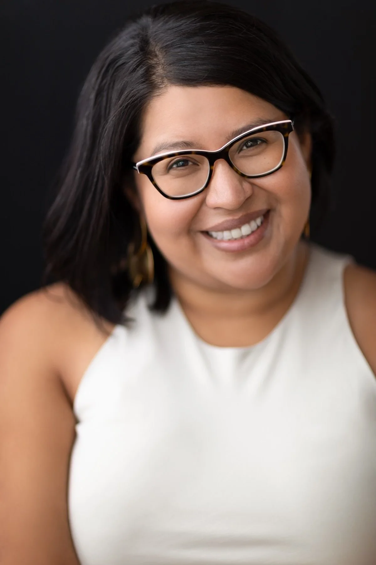 Close-up portrait of a woman with glasses smiling and looking at the camera against a dark background.