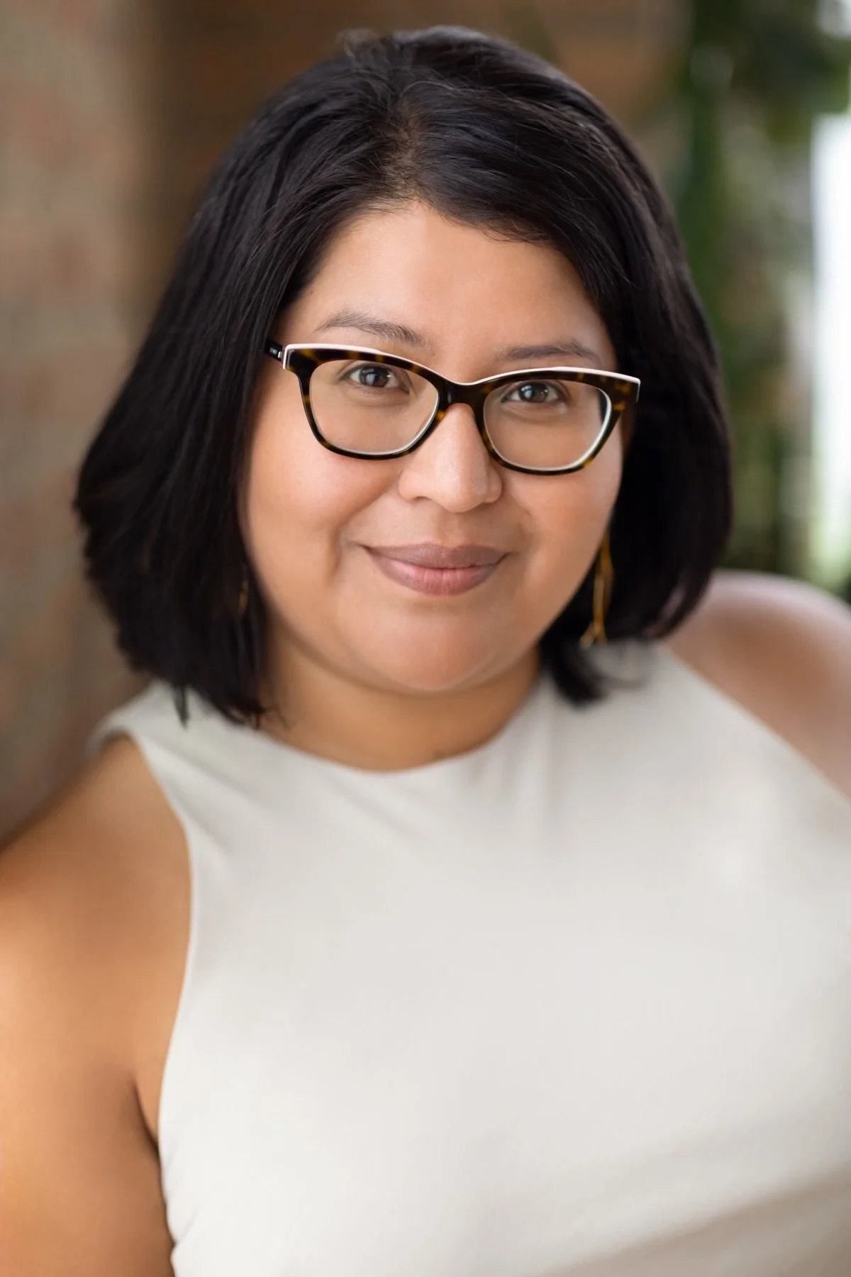 A woman with shoulder-length black hair, wearing glasses with tortoiseshell frames, smiling, dressed in a sleeveless white top, with a blurred green background.