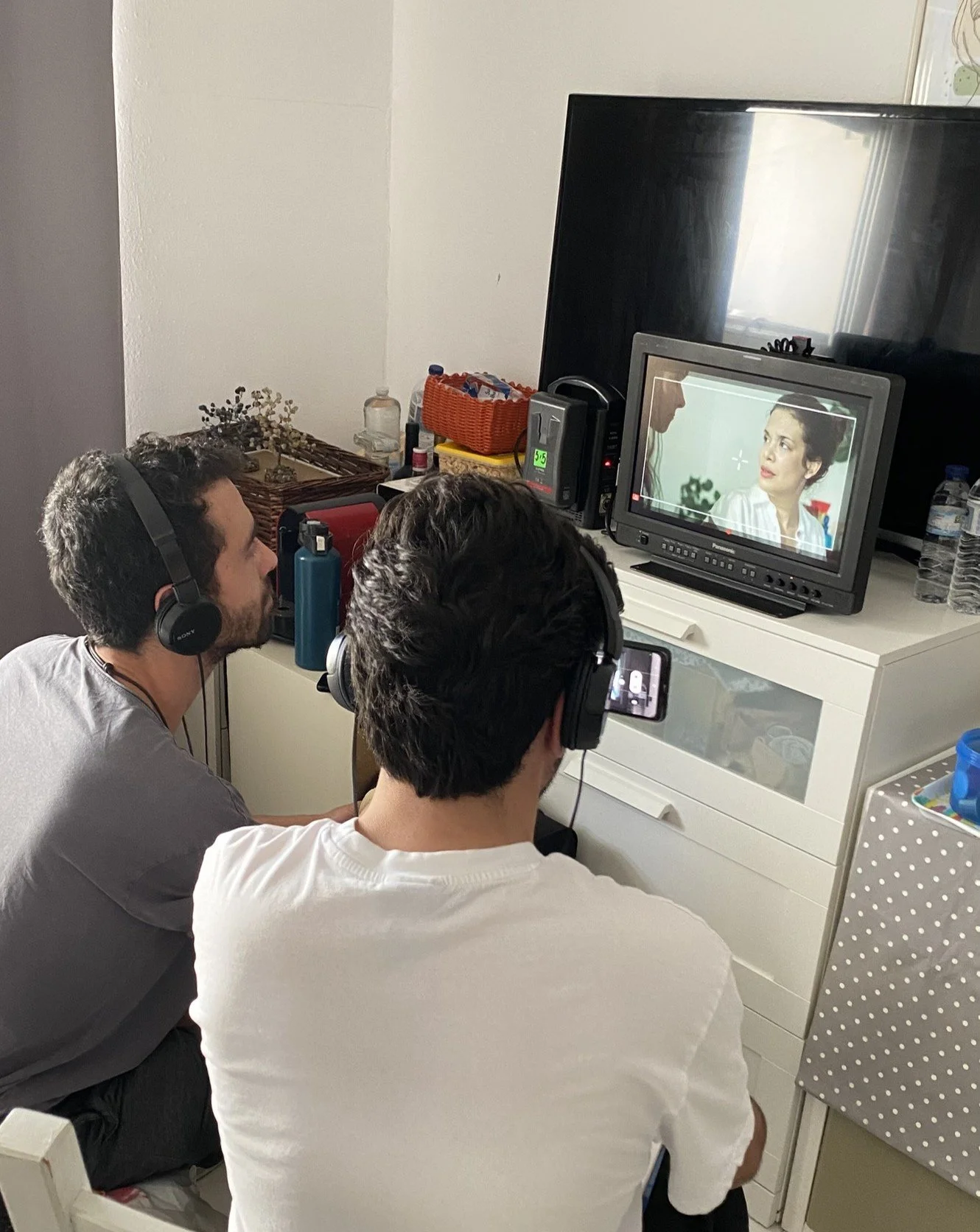 Duas pessoas assistindo uma cena de televisão em um ambiente interno, usando headphones, possivelmente gravando ou produzindo conteúdo relacionado ao programa exibido.