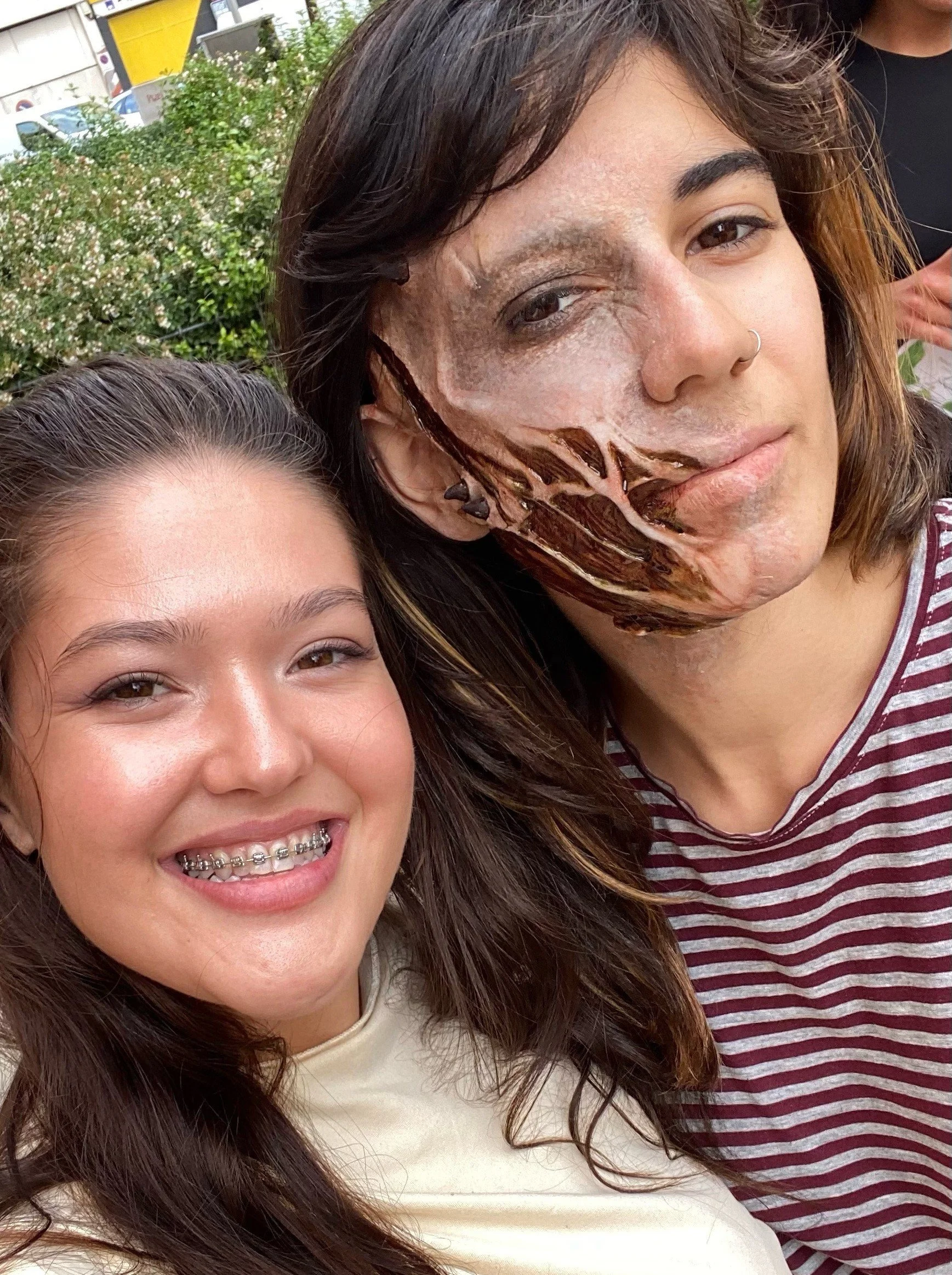 Dupla de jovens sorrindo para a câmera, uma delas com aparelho nos dentes, outra com maquiagem artística de couro na face, ao ar livre com vegetação ao fundo.