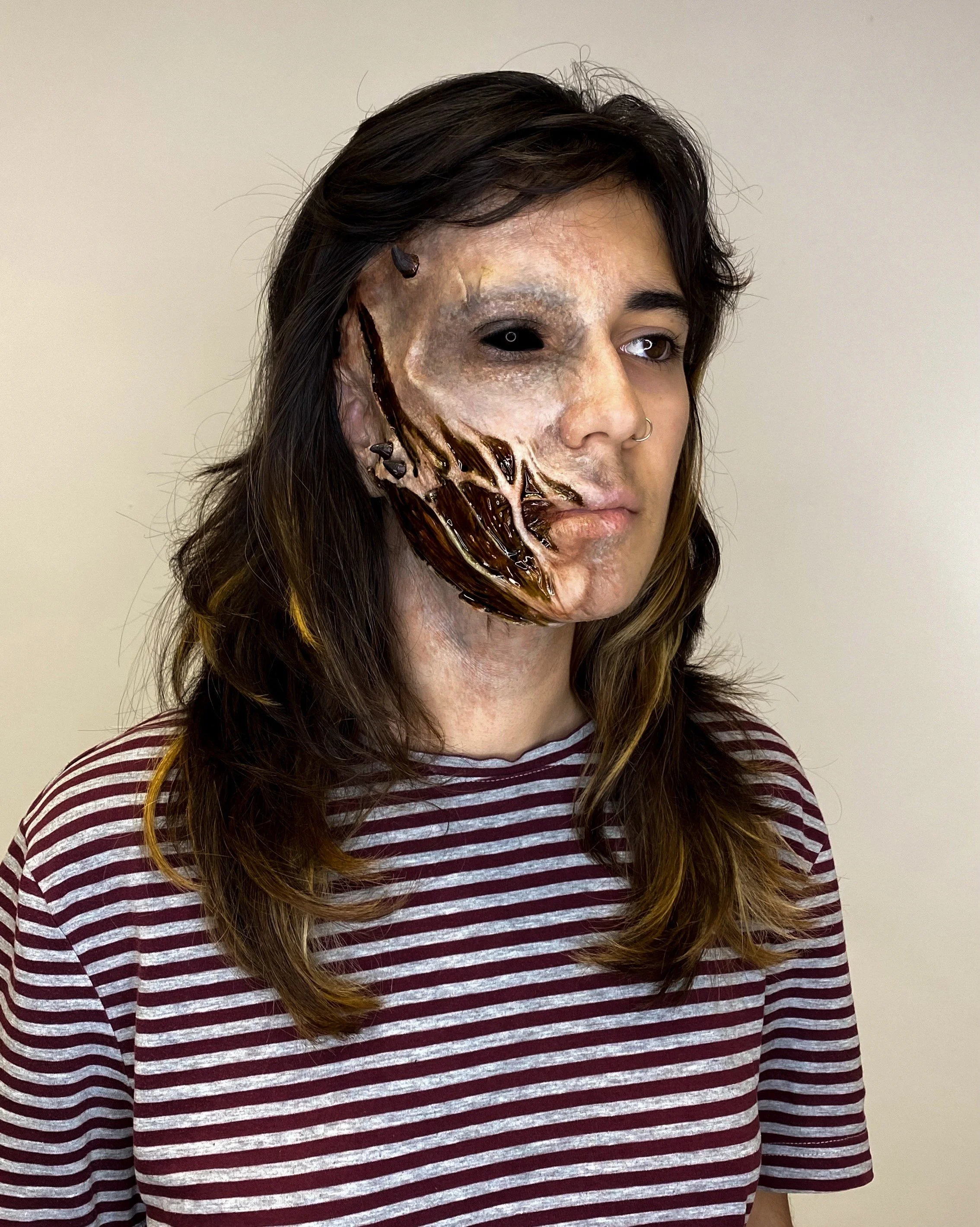 Homem com maquiagem de rosto assustador, metade do rosto é uma caveira escura, com detalhes de ossos e uma expressão macabra, usando uma blusa listrada vermelha e branca.