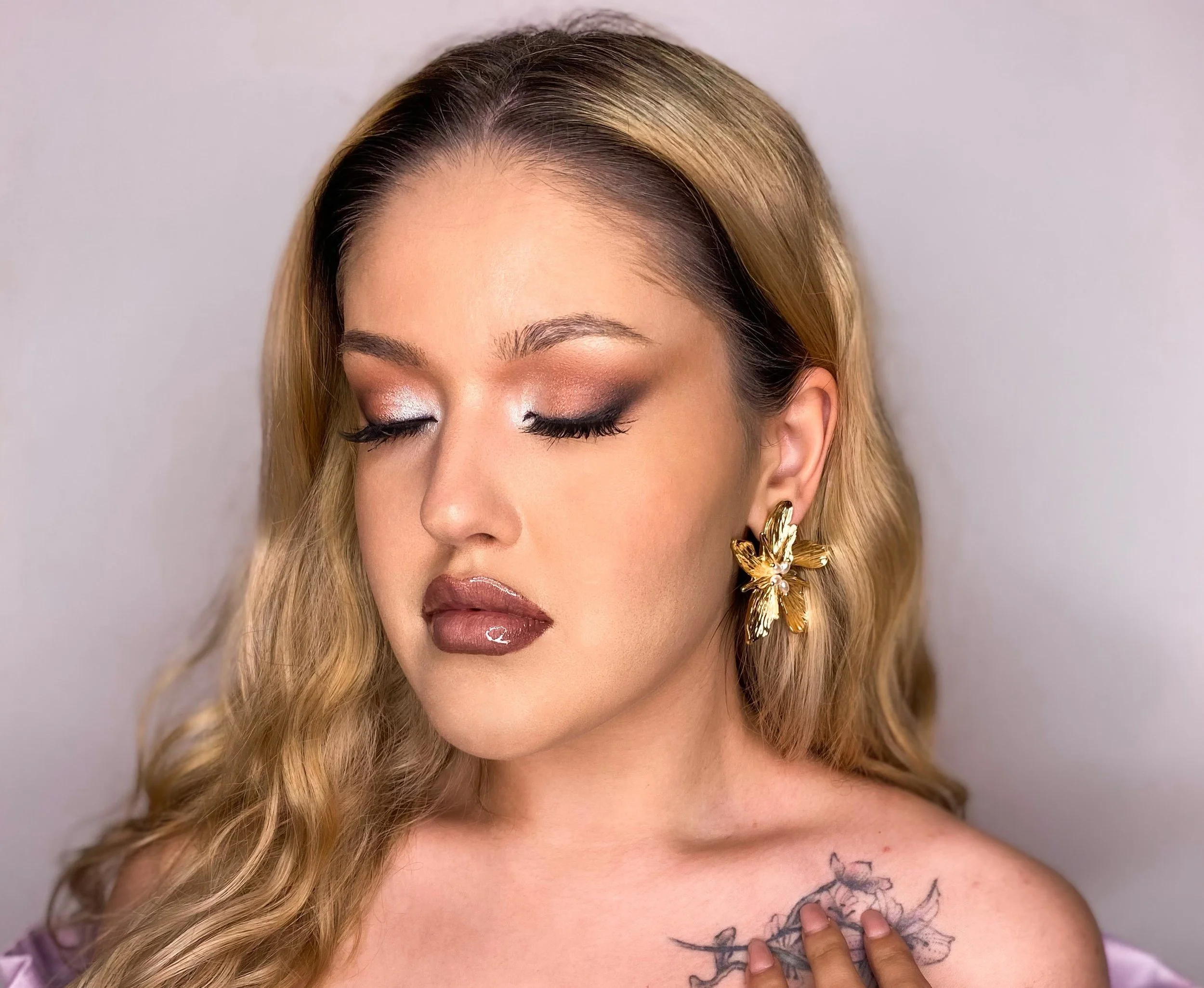 mulher com cabelo liso loiro, maquiagem com olhos fechados, lábios brilhantes, usando brincos dourados em forma de borboleta, tocando um tatuagem no ombro.