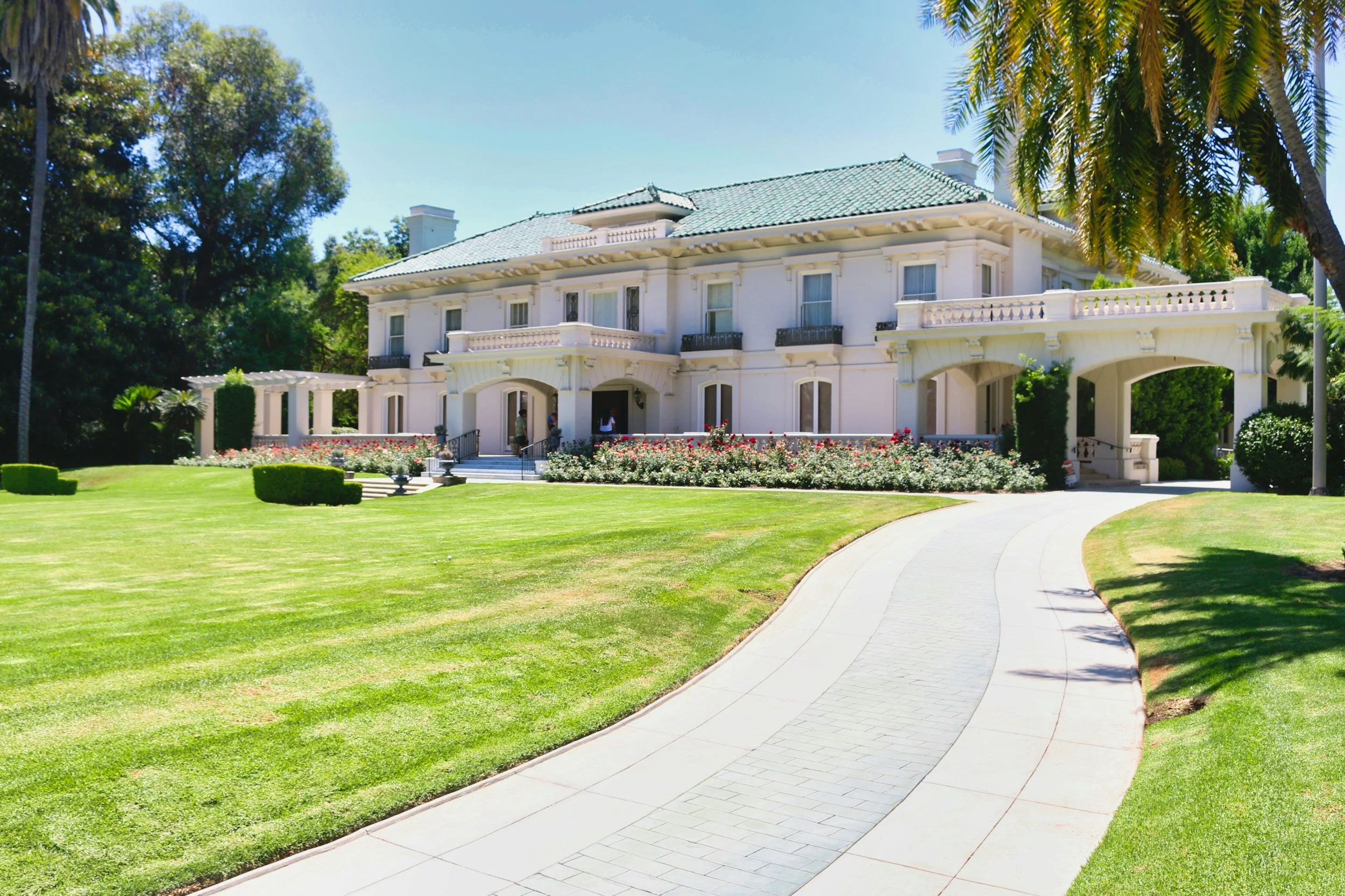 A large, white, multi-story San Diego area mansion with a green tiled roof, surrounded by a well-maintained lawn and flower beds. There is a winding paved walkway leading to the entrance, with palm trees and other greenery around.