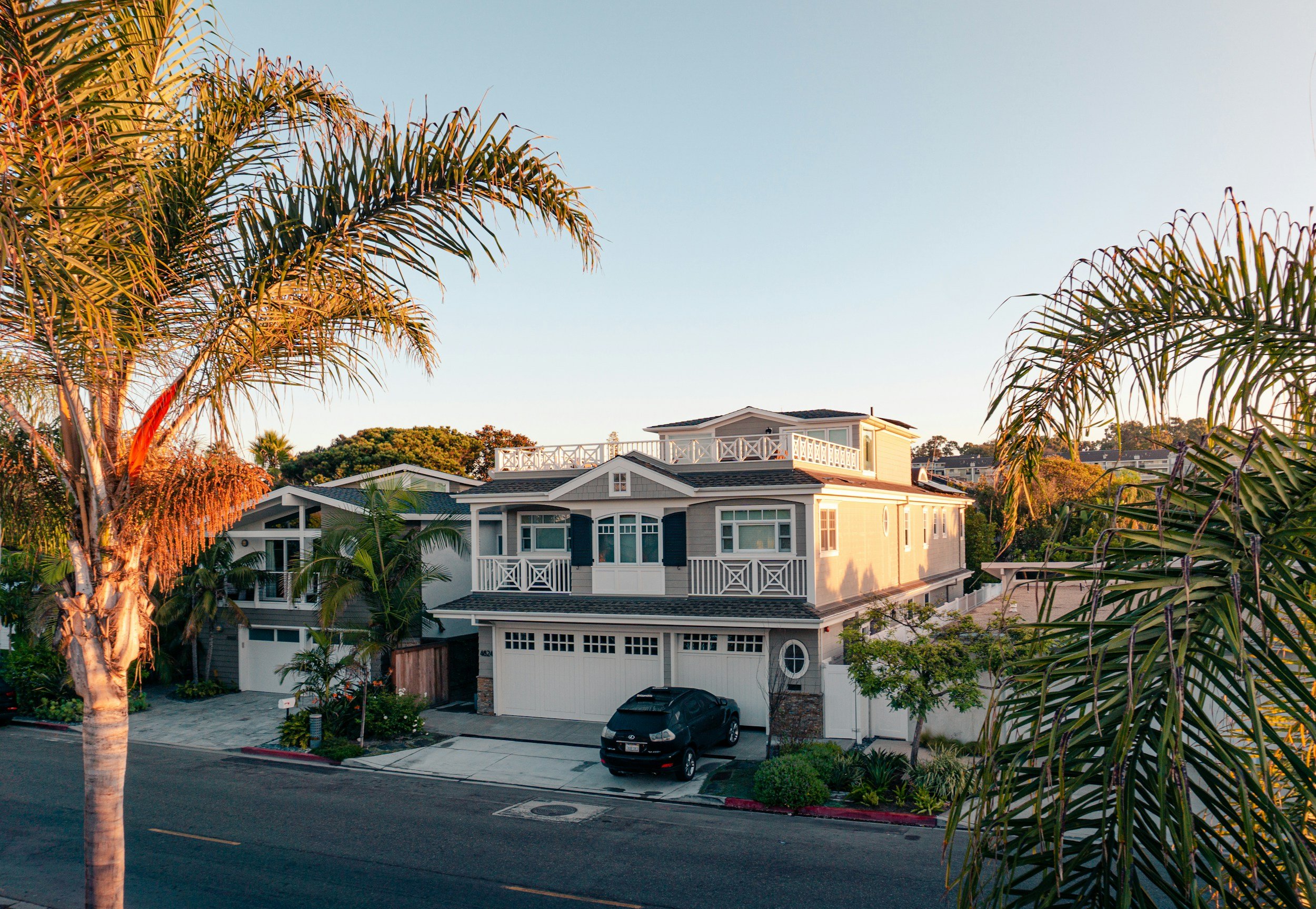A modern two-story San Diego house with a white garage door, a black car parked in front, and lush palm trees surrounding the property during sunset