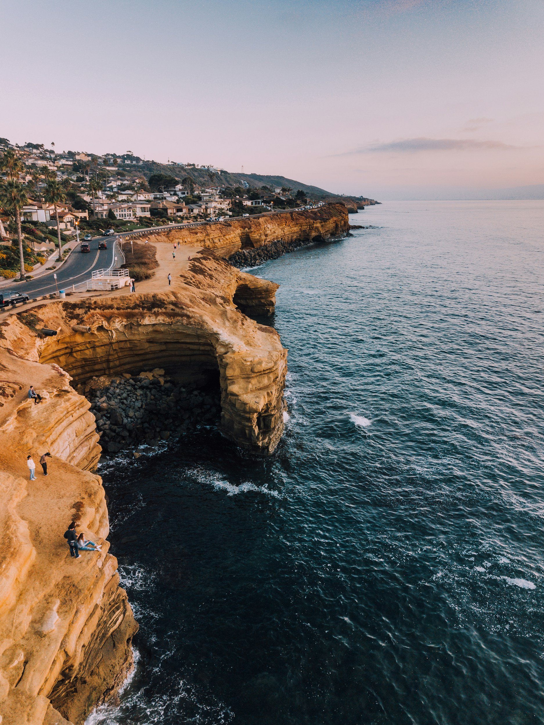 San Diego Cliffside along the coast with sandy formations, people walking and sitting, and residential houses on a hillside above the shoreline during sunset.