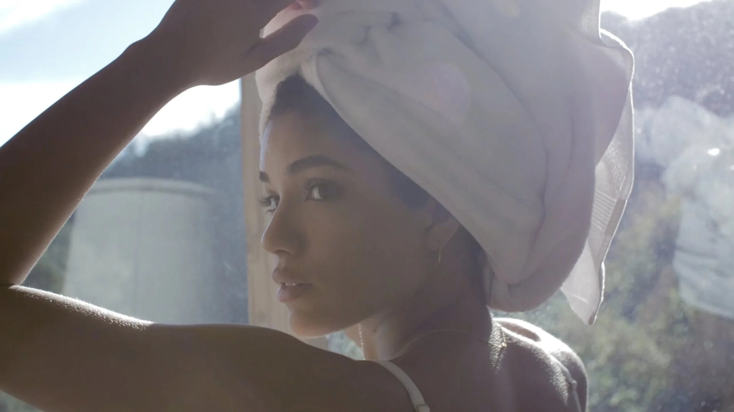 Woman with a towel wrapped around her head, looking out a window with sunlight shining through.