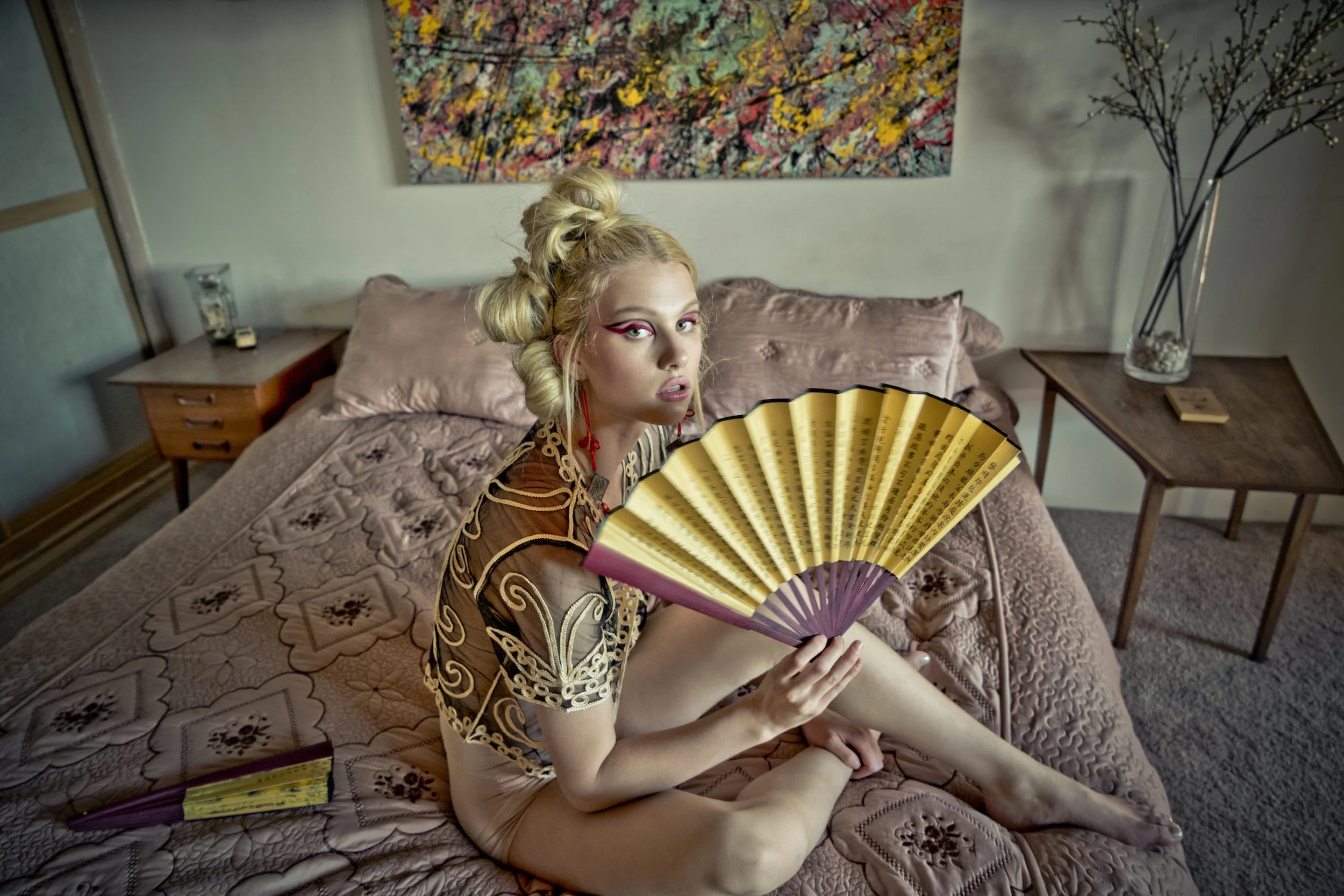 Young woman with blonde hair styled in updo, sitting on a bed with pink bedding, holding a large decorative fan, wearing a sheer patterned top, in a bedroom with artwork on the wall and side tables.