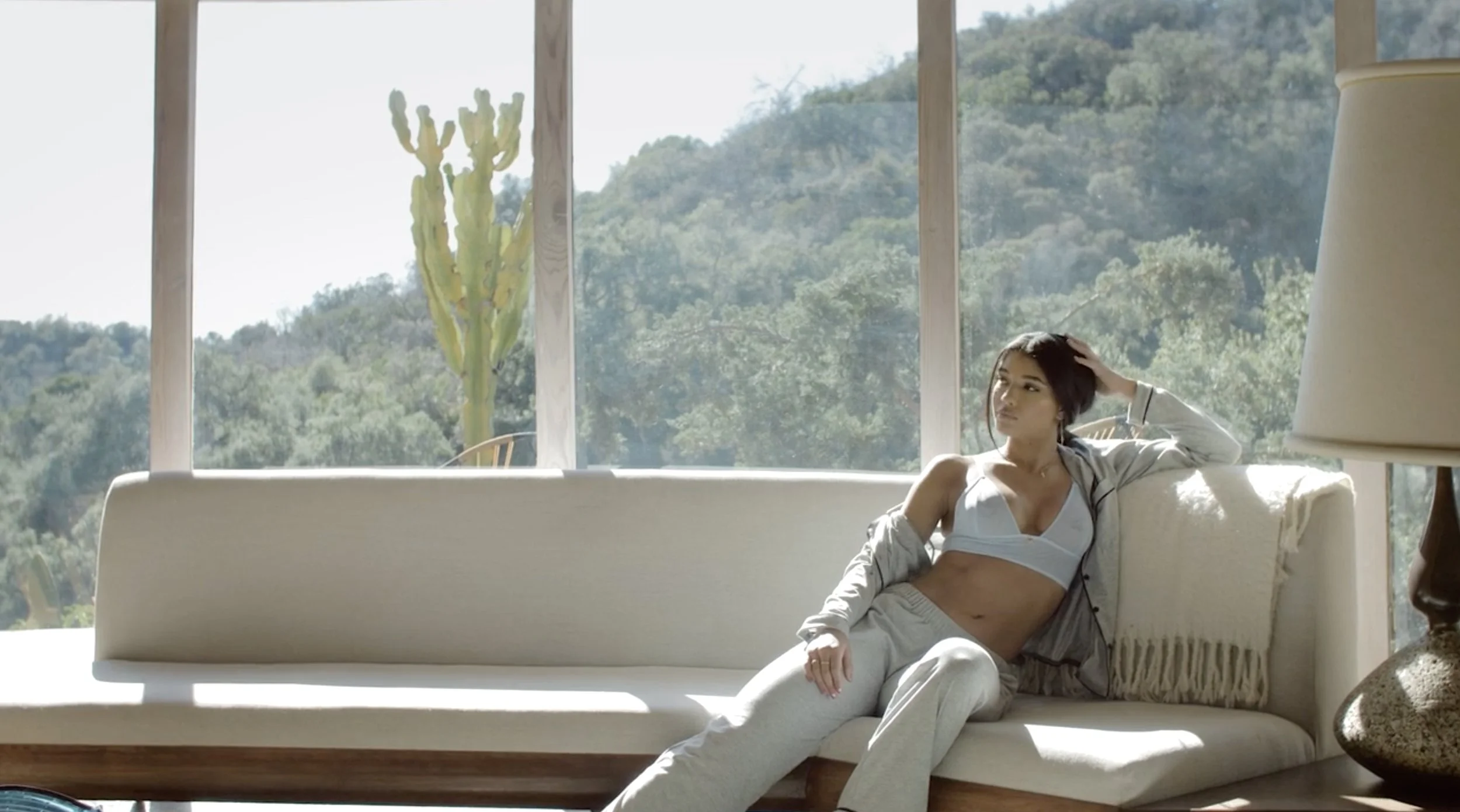 Young woman sitting on a cream-colored sofa in front of large windows with a scenic view of green hills and a cactus outside. She has dark hair and is wearing a sports bra, sweatpants, and an open jacket, with a relaxed pose and one hand resting on h