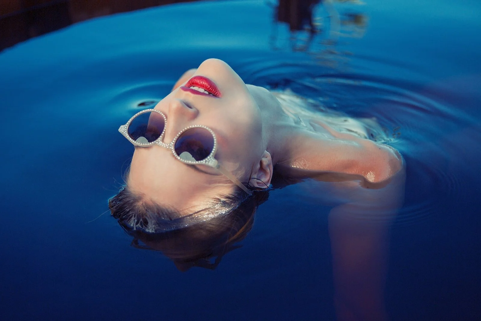 A woman floating on her back in a pool, wearing sunglasses with pearl-like decorations and red lipstick.