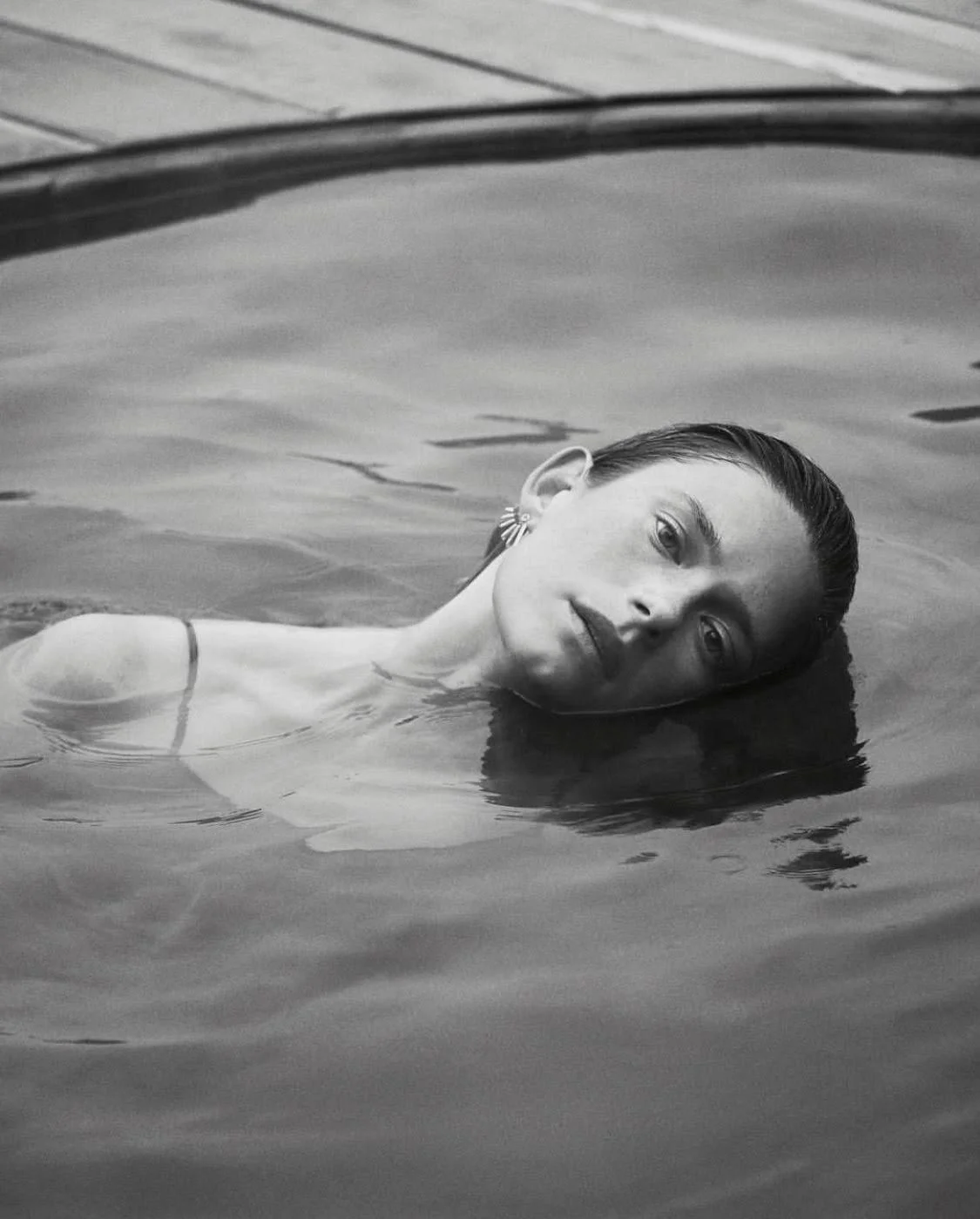 A woman with slicked-back hair and earrings is floating in a swimming pool, resting her head on the water, with a calm expression.
