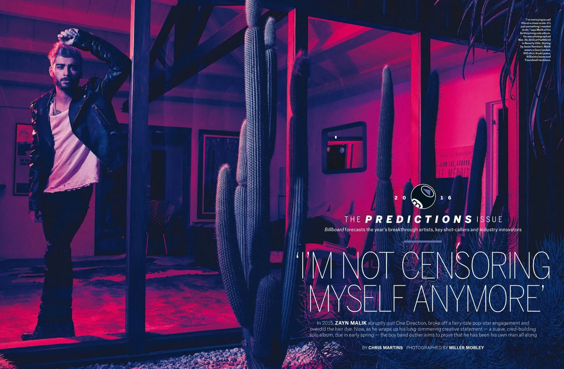 A man with styled hair and a beard, wearing a black leather jacket over a white torn shirt, poses indoors next to a large cactus plant. The room has pink and purple lighting, with framed artwork on the walls and a patterned rug on the floor.