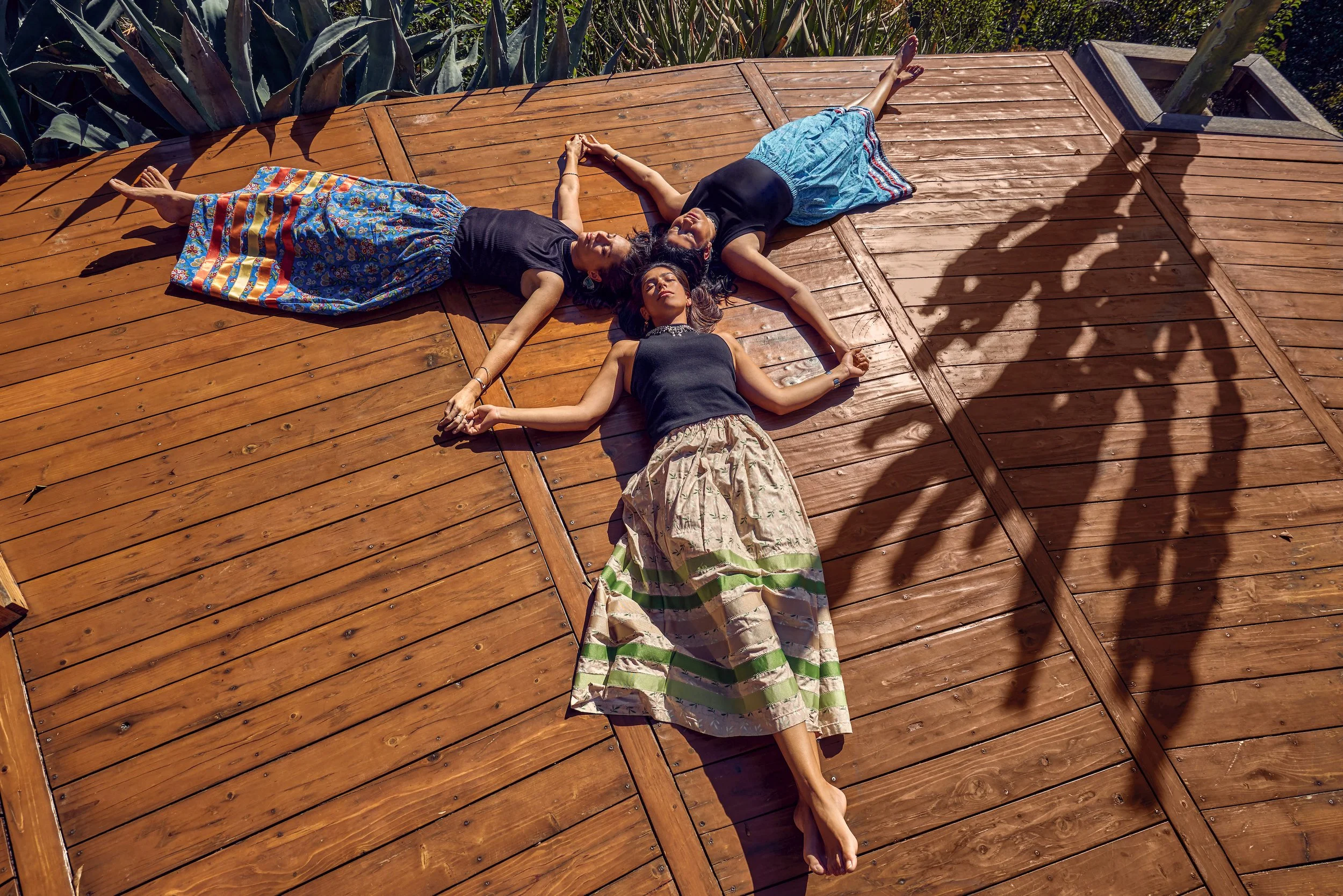 Three women lying on a wooden deck holding hands in a star shape, sunlight casting shadows, surrounded by greenery.