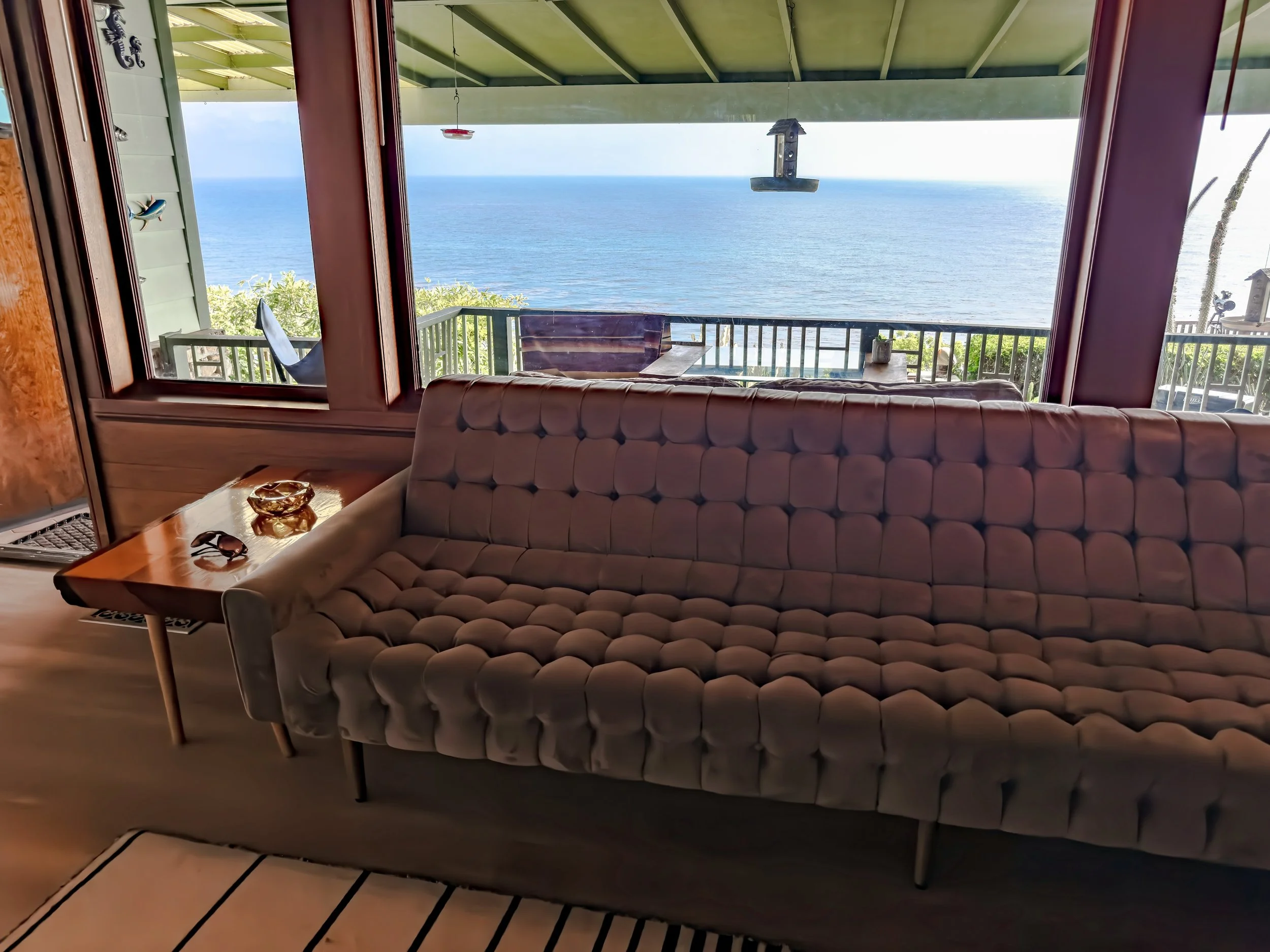 Interior view of a living room with a large window overlooking the ocean, showing a balcony with outdoor furniture and a birdhouse hanging from the ceiling.