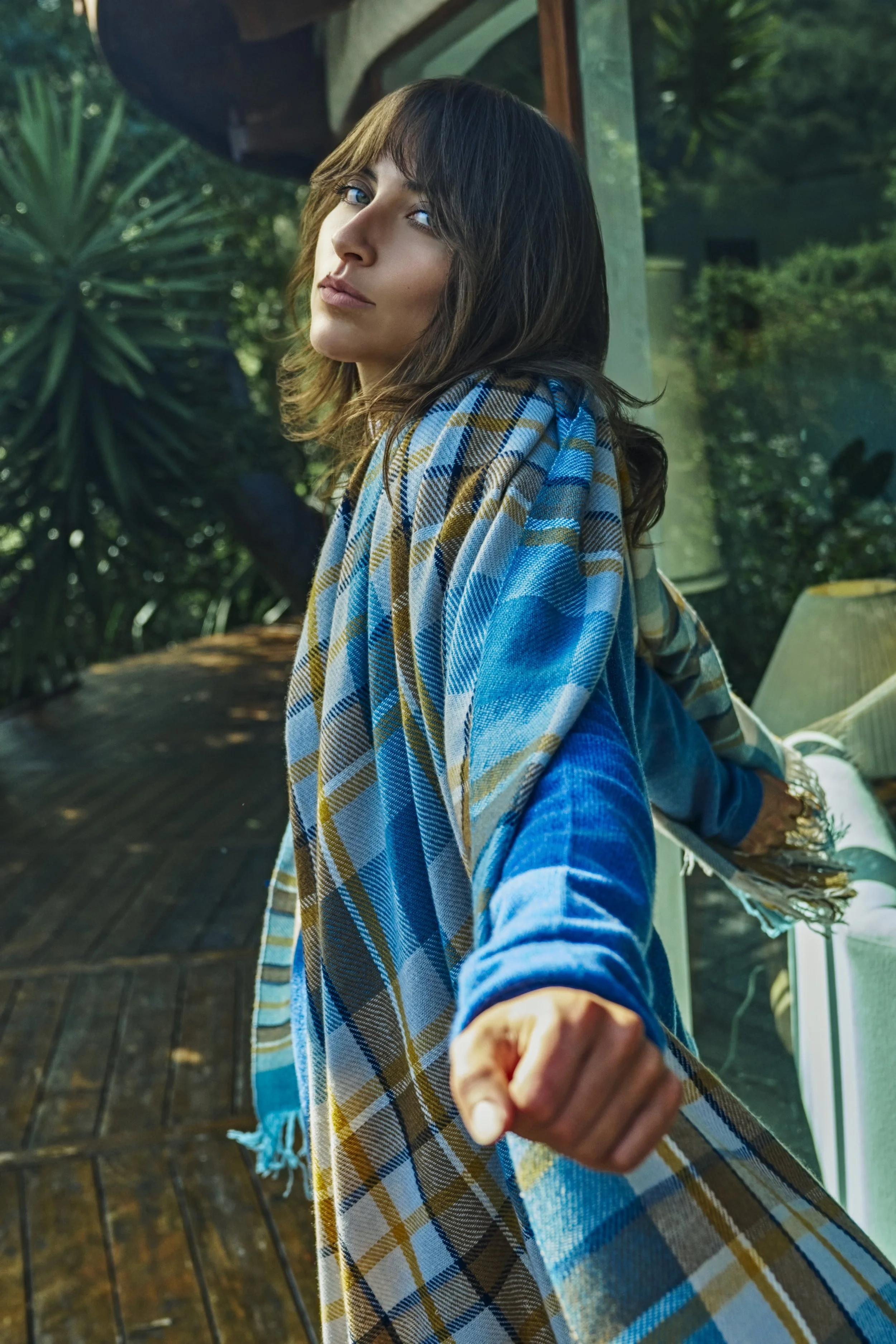 Young woman taking a selfie on a wooden deck with lush greenery in the background, wearing a blue plaid scarf and blue shirt.