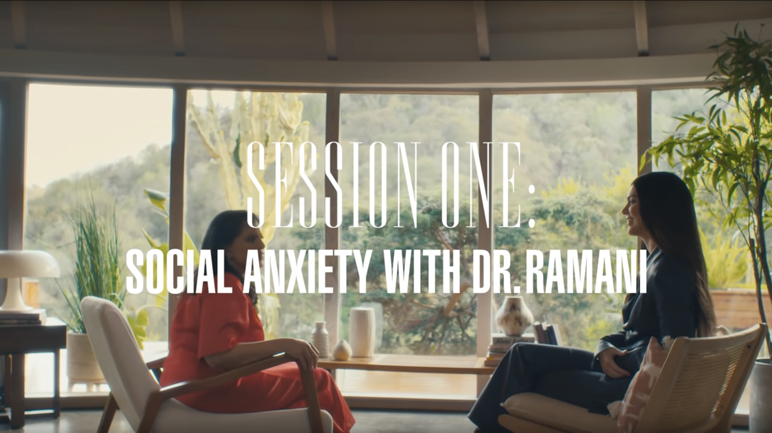 Two women sit facing each other in a comfortable room with large windows overlooking a lush outdoor landscape. One woman wears a red dress, and the other wears a black blazer. The scene is set for a discussion, with text overlay reading 'Session One: