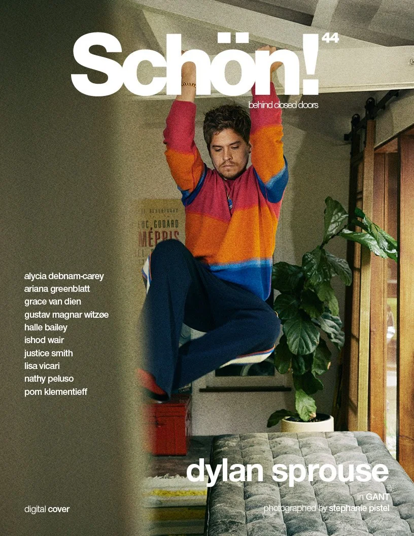 A young man with dark, curly hair and a beard is hanging upside down in a room, holding onto a wooden beam. He is wearing a colorful striped sweater with red, orange, blue, and purple and dark pants. Behind him is a large green plant with broad leave