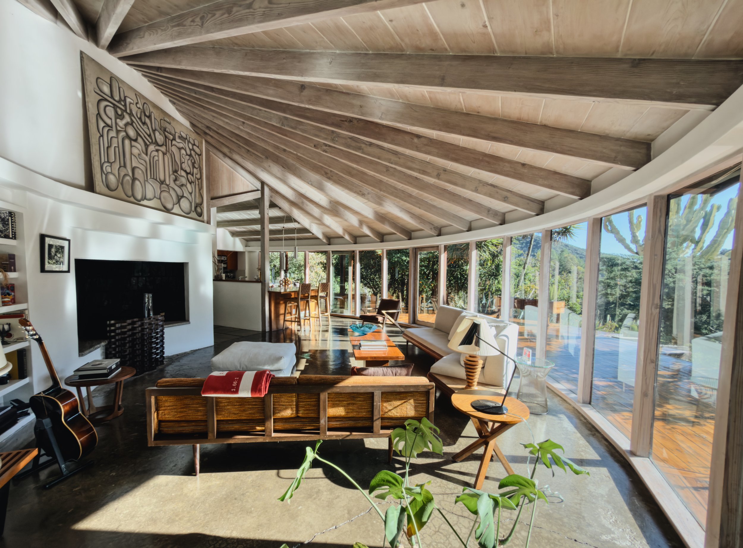 Spacious living room with a large curved wall of floor-to-ceiling windows overlooking greenery, wooden ceiling with exposed beams, white walls, modern furniture including sofas and chairs, a guitar, and decorative items.