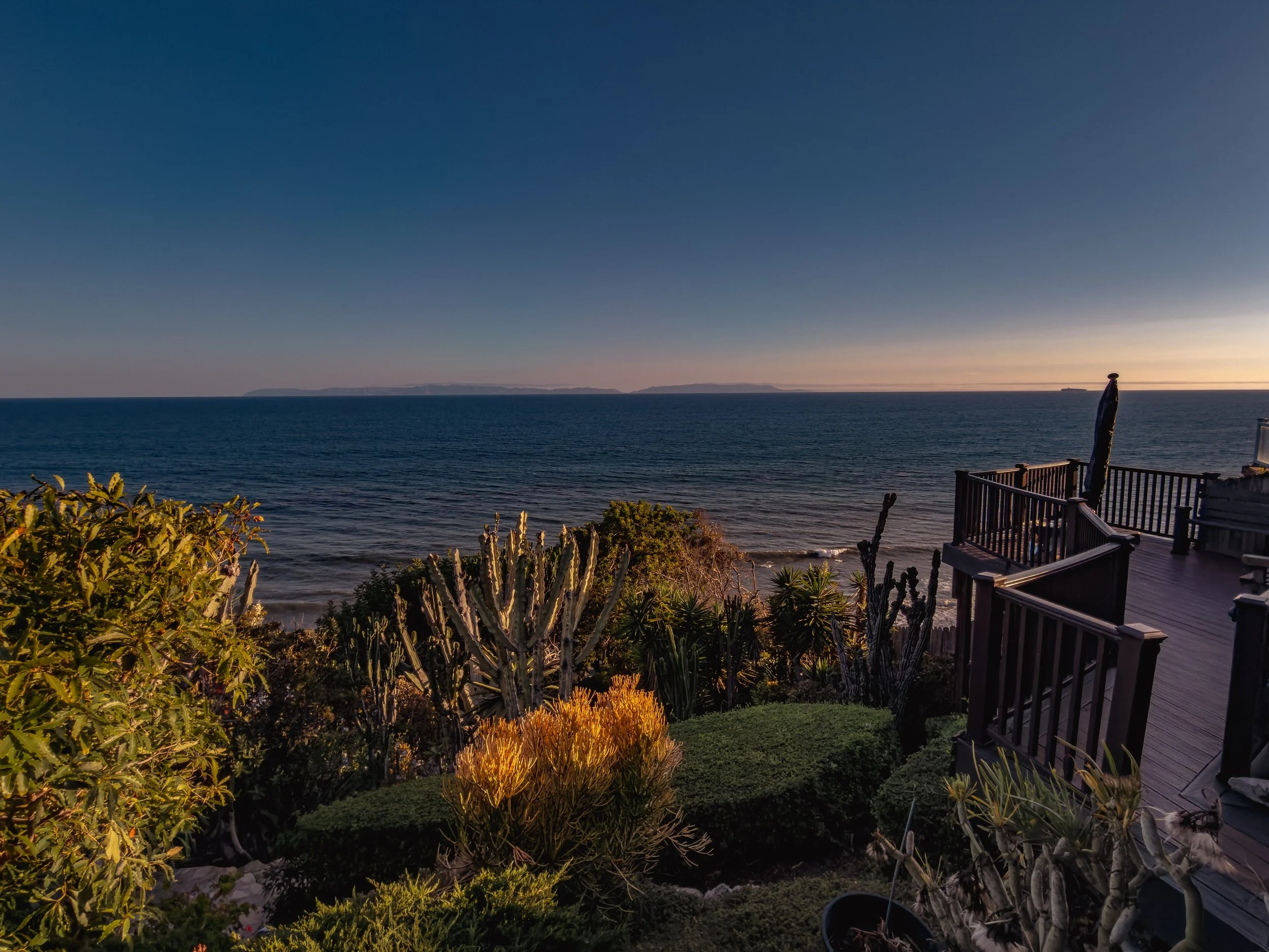 A view of a calm ocean or sea during sunset with distant landforms on the horizon. In the foreground, there is a wooden deck with a railing, surrounded by various green desert plants and cacti. A wrapped umbrella is visible on the deck.