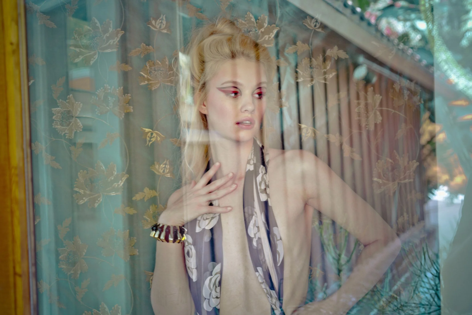 A young woman with blonde hair styled in an updo, wearing dramatic eye makeup and a floral halter top, gazes through a window with floral-patterned curtains, touching her neck with her left hand and resting her right arm on her hip.