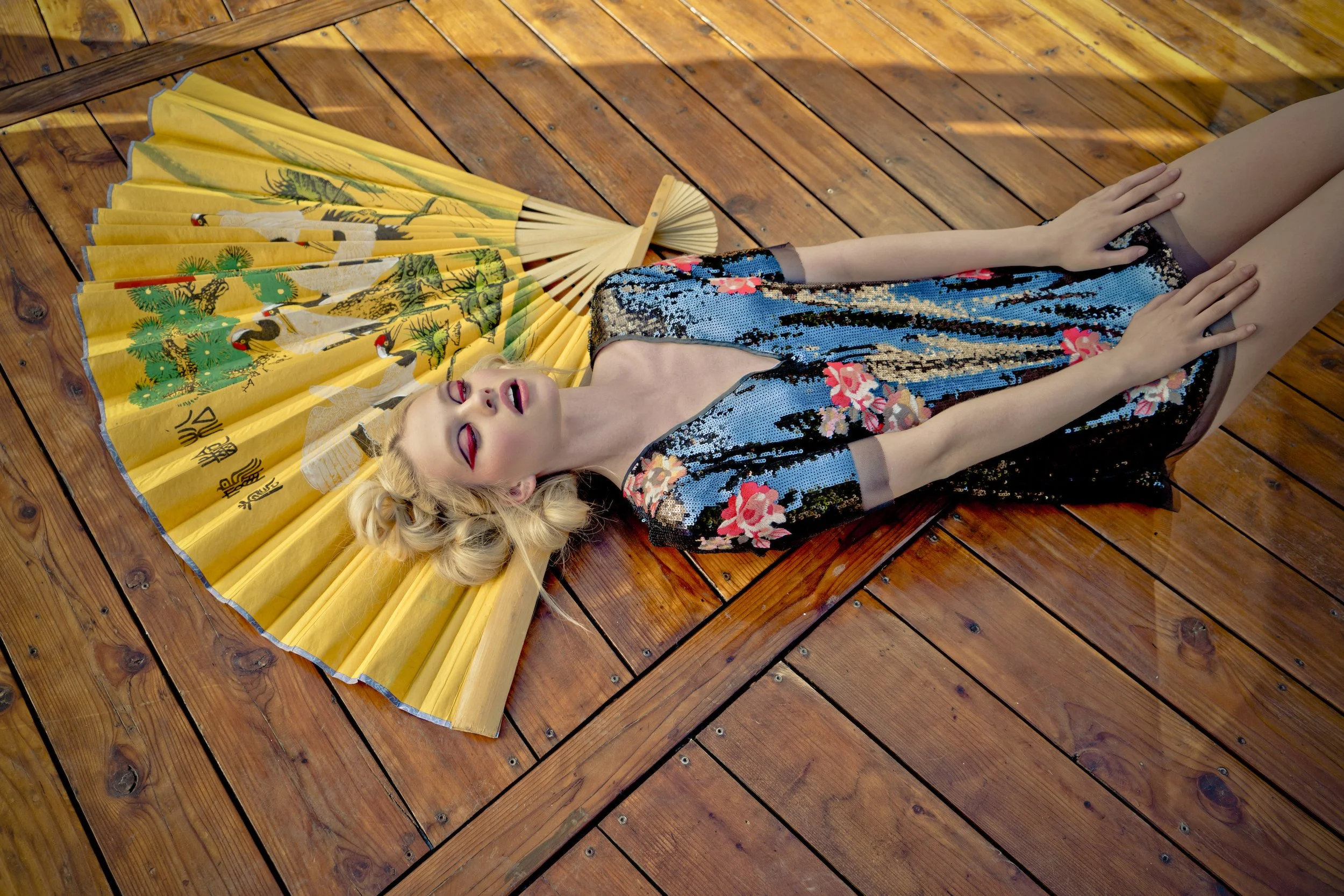 A woman with blonde hair lying on a wooden floor next to a yellow decorative fan with Asian art and writing, wearing a sequined dress with floral prints, her eyes closed and head tilted back.